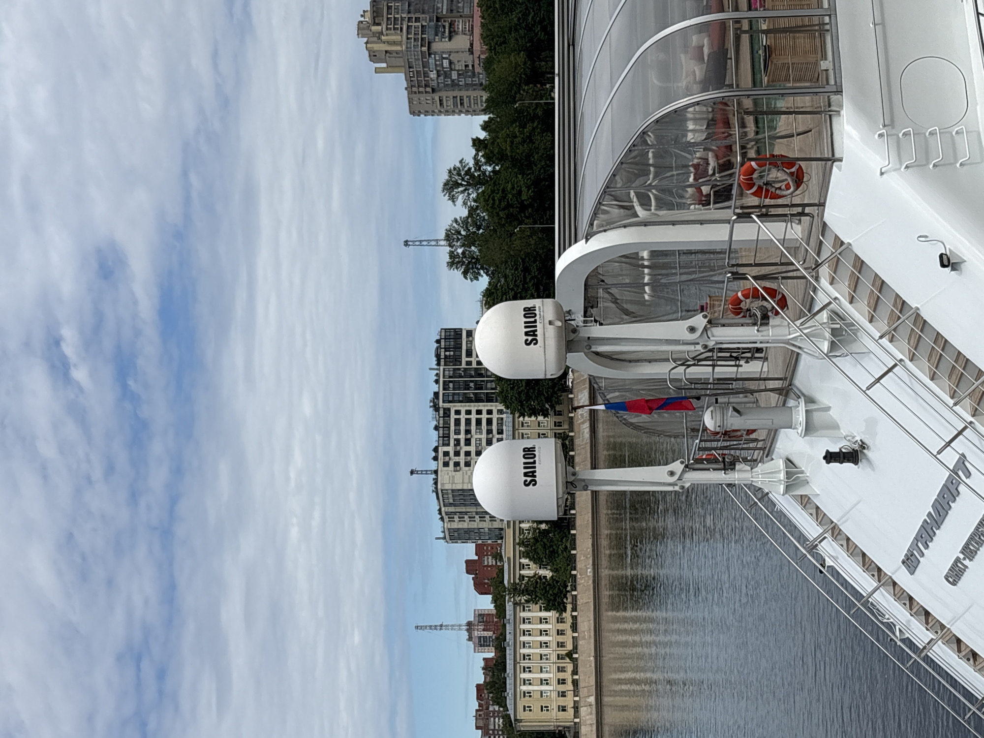 The image depicts a view from the deck of a white yacht, likely docked in Saint Petersburg, Russia. The yacht's deck is visible in the foreground, with polished metal railings and stairs leading down. Two white satellite domes, branded with "SAILOR", are prominently positioned on the deck. A small Russian flag flies from a pole. Behind the yacht, a body of water, possibly a river or canal, reflects the sky and the city.
The background features a cityscape with several buildings of varying architectural styles. There are older, institutional-looking buildings with multiple windows, and more modern, taller apartment blocks. Green trees are interspersed between the structures. The sky is partly cloudy, with a mix of white and gray clouds against a pale blue backdrop. The overall atmosphere is calm and suggests a day with moderate weather conditions. The yacht itself appears to be a luxurious vessel, indicated by its clean white finish and the presence of amenities visible under a transparent canopy, such as seating and what appears to be a small pool. The inscription on the yacht's hull reads "ШТАНДАРТ" and "САНКТ-ПЕТЕРБУРГ", confirming its location and name, which translates to "Standard" and "Saint Petersburg".