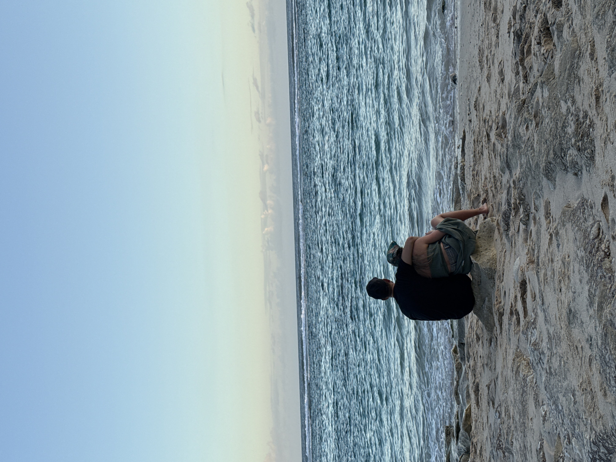 The image shows a beach scene in Ungasan, Indonesia. A man in a black shirt sits on the sand near the water's edge. A child rests on his lap. The sky is light blue, transitioning to a light yellow near the horizon. The ocean has a textured, wavy surface. The beach consists of light-colored sand.