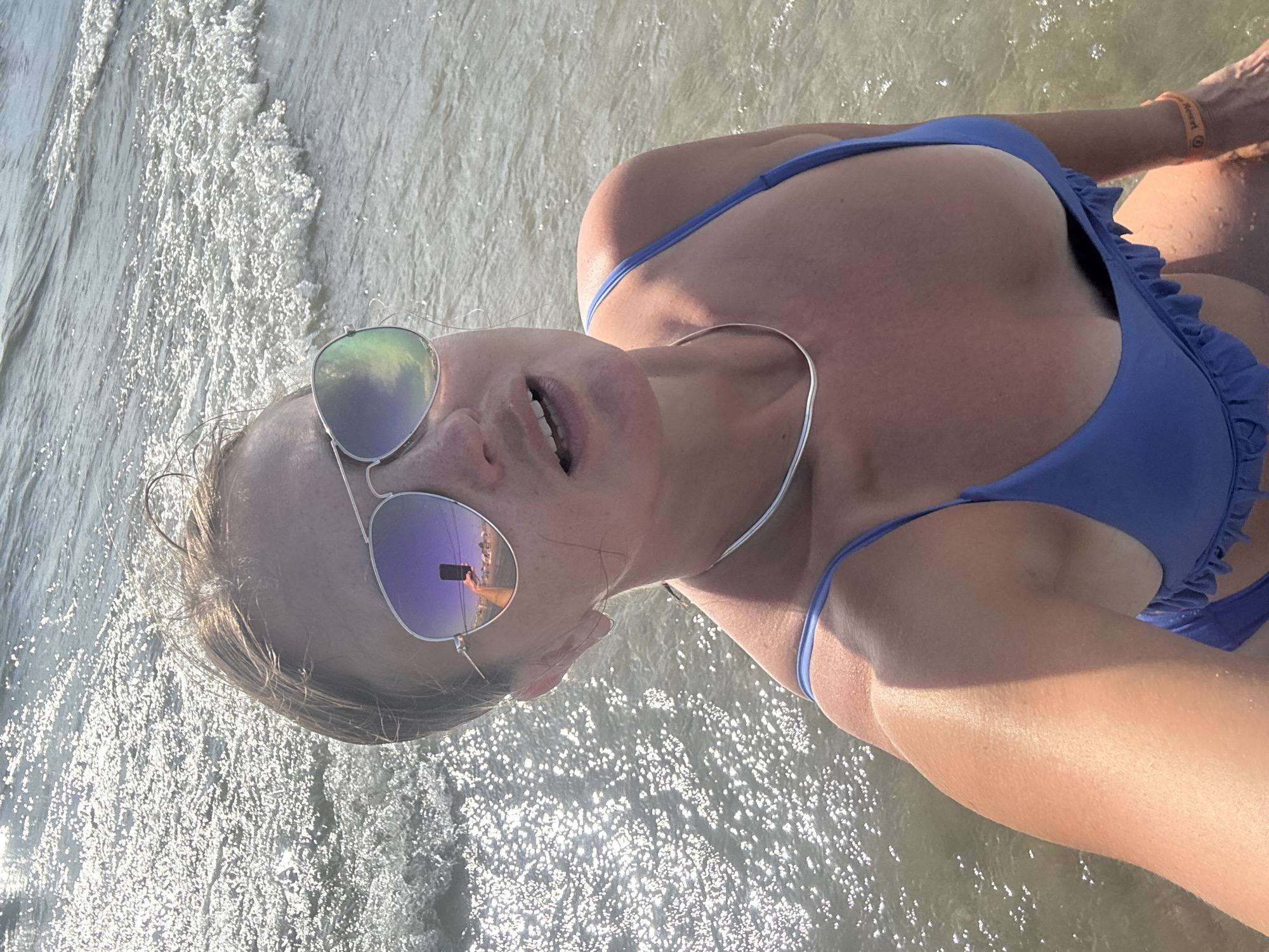 A woman, seen from the chest up, is posing for a selfie in shallow water at the beach in Le Grau-du-Roi, France. She has short brown hair, is wearing aviator sunglasses, and a blue bikini with ruffled trim. Her right arm is extended, holding the camera, and she is wearing an orange wristband. The water is clear with a sandy bottom, and sunlight glints off the surface. The background shows gentle waves and the sea.