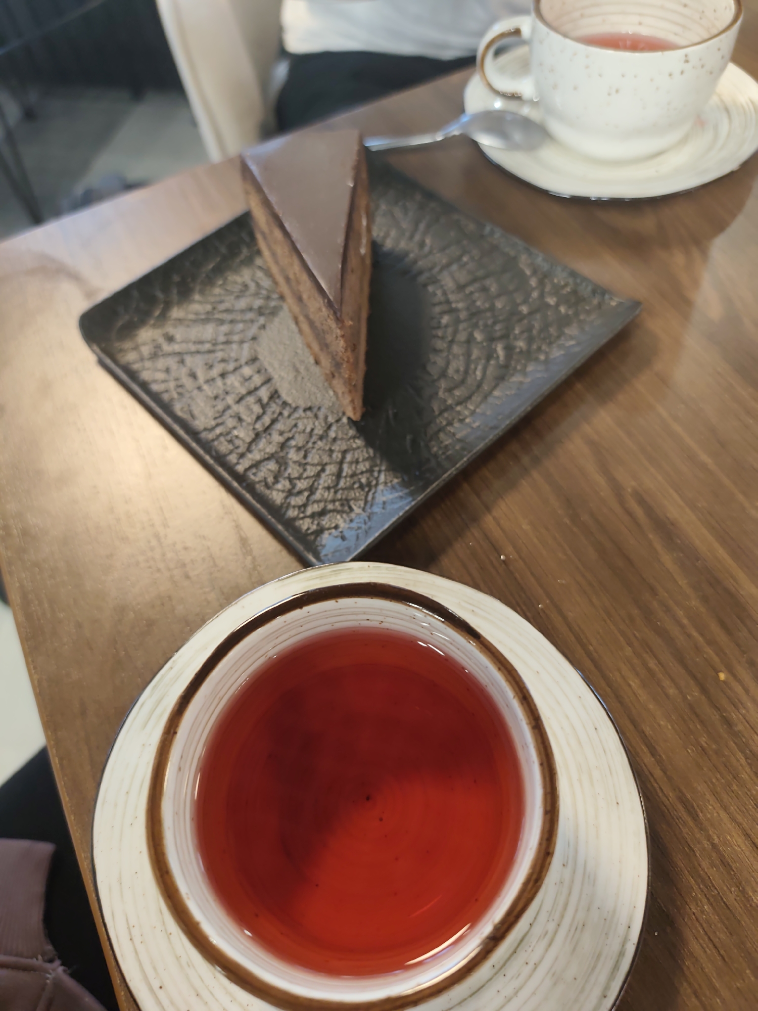 The image presents a high-angle view of a wooden table, likely in a cafe or restaurant setting in Chaikovsky, Russia. The primary subjects are two cups of tea and a slice of chocolate cake.
In the foreground, a white and brown ceramic teacup, resting on a matching saucer with a subtle concentric circle pattern, holds a vibrant reddish-orange liquid, possibly a fruit or berry tea. The cup's rim has a distinctive brown band. Further back and to the right, another white ceramic teacup with a speckled design is visible on its saucer, containing a lighter red beverage. A silver spoon is placed on this saucer.
Between the two tea settings, a triangular slice of dark chocolate cake is displayed on a square, dark gray or black plate with a distinct textured, abstract pattern. The cake features a smooth, glossy dark chocolate glaze on top and shows visible layers of chocolate sponge and filling along its side, suggesting a rich dessert like a Sacher Torte.
A person's arm, dressed in dark clothing, is partially visible in the out-of-focus background, implying someone is seated at the table. The scene depicts a quiet moment of enjoying beverages and dessert. No text is visible in the image.
