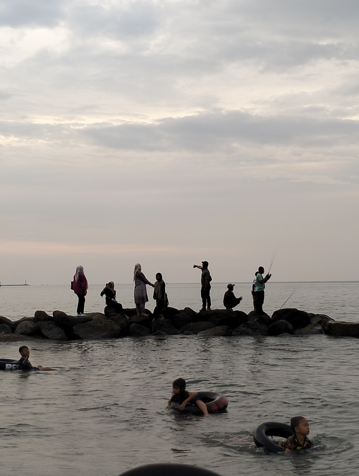 A coastal scene in Lhokseumawe, Indonesia, depicts a tranquil, overcast environment at an unspecified time, likely dawn or dusk, given the diffused light. The horizon line is visible across the calm, grey-blue sea, with a faint structure resembling a lighthouse or pole on the far left. A rocky breakwater extends across the mid-ground, partially submerged.
Ten individuals are present on the breakwater. On the left section, five individuals, primarily appearing to be females wearing head coverings (hijabs), are distributed: one stands in a pink top, one sits facing right, one stands in a light-colored dress, and two others are partially visible standing or crouching. Their actions include standing, sitting, and potentially interacting. On the right section of the breakwater, five individuals, appearing to be males, are present: one stands pointing to the right, one squats, two are actively engaged in fishing with rods, and one is partially obscured near the water's edge.
In the immediate foreground, three children are in the calm water. One child on the far left and one on the right are floating in black inner tubes. A third child in the center-foreground floats in a dark reddish-black inner tube. The children are either looking towards the right or forward. The water exhibits minor ripples.