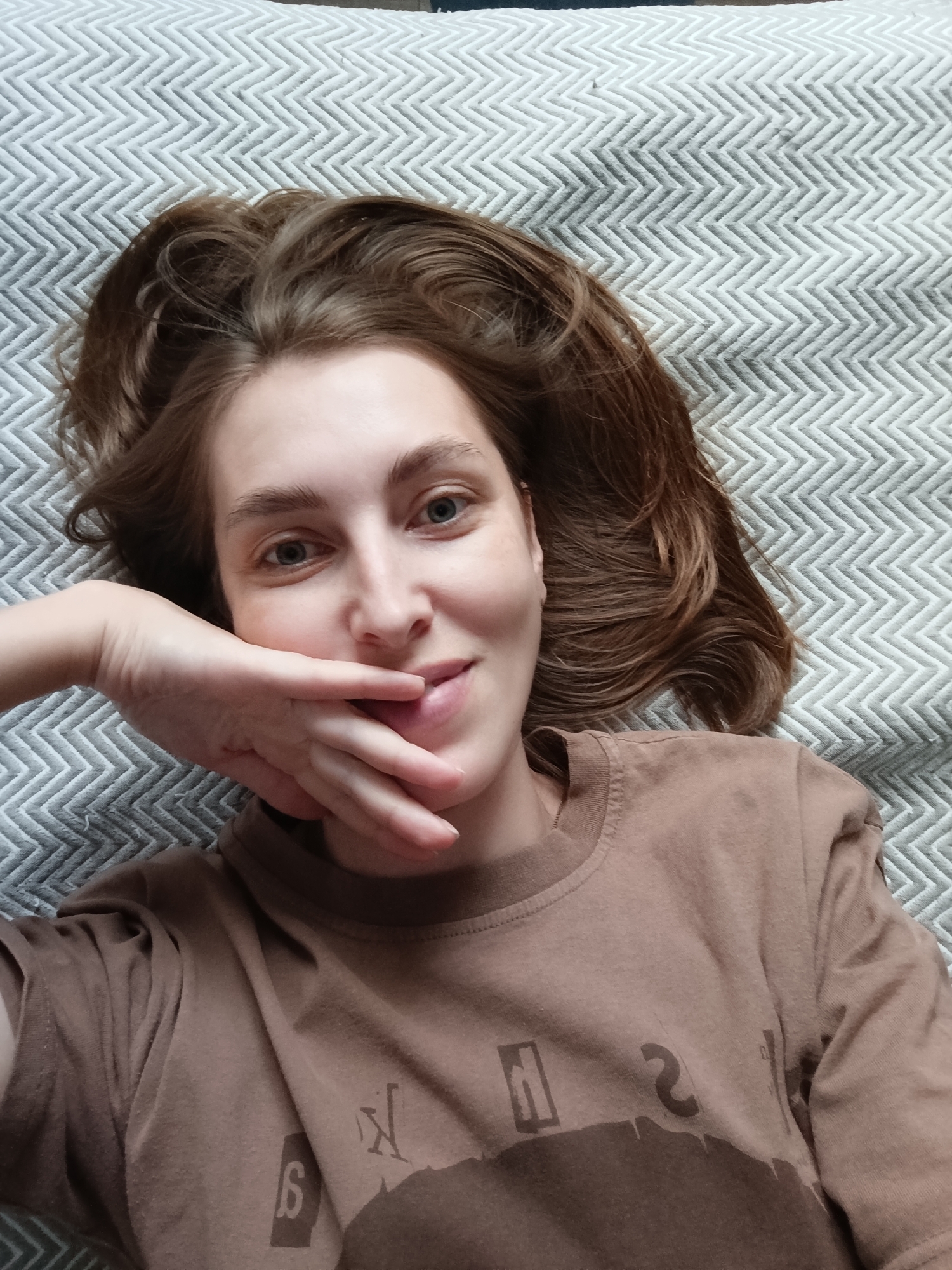 A young woman with brown hair lies on her back on a textured grey surface, looking at the camera. Her right hand is raised to her face, with her fingers covering her mouth and nose. She is wearing a brown t-shirt with a faded graphic design on the front. The background is a grey, chevron-patterned blanket or comforter. The image appears to be taken indoors in a domestic setting, possibly a bedroom. There is no information in the image to suggest a location in Chaikovsky, Russia.