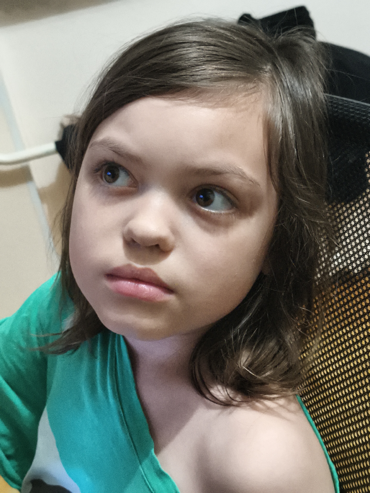 The image is a close-up, eye-level, medium shot of a young girl with medium-length brown hair and brown eyes. She is wearing a teal-colored shirt. Her head is turned to her left, and she is looking upwards and to her left. Her expression appears to be serious or contemplative. The background is out of focus and appears to be an indoor setting with a wall and a textured surface that resembles the back of a chair. The lighting suggests an indoor environment, possibly with artificial light. The location in Chaikovsky, Russia, is not discernible from the visual information provided in the image.