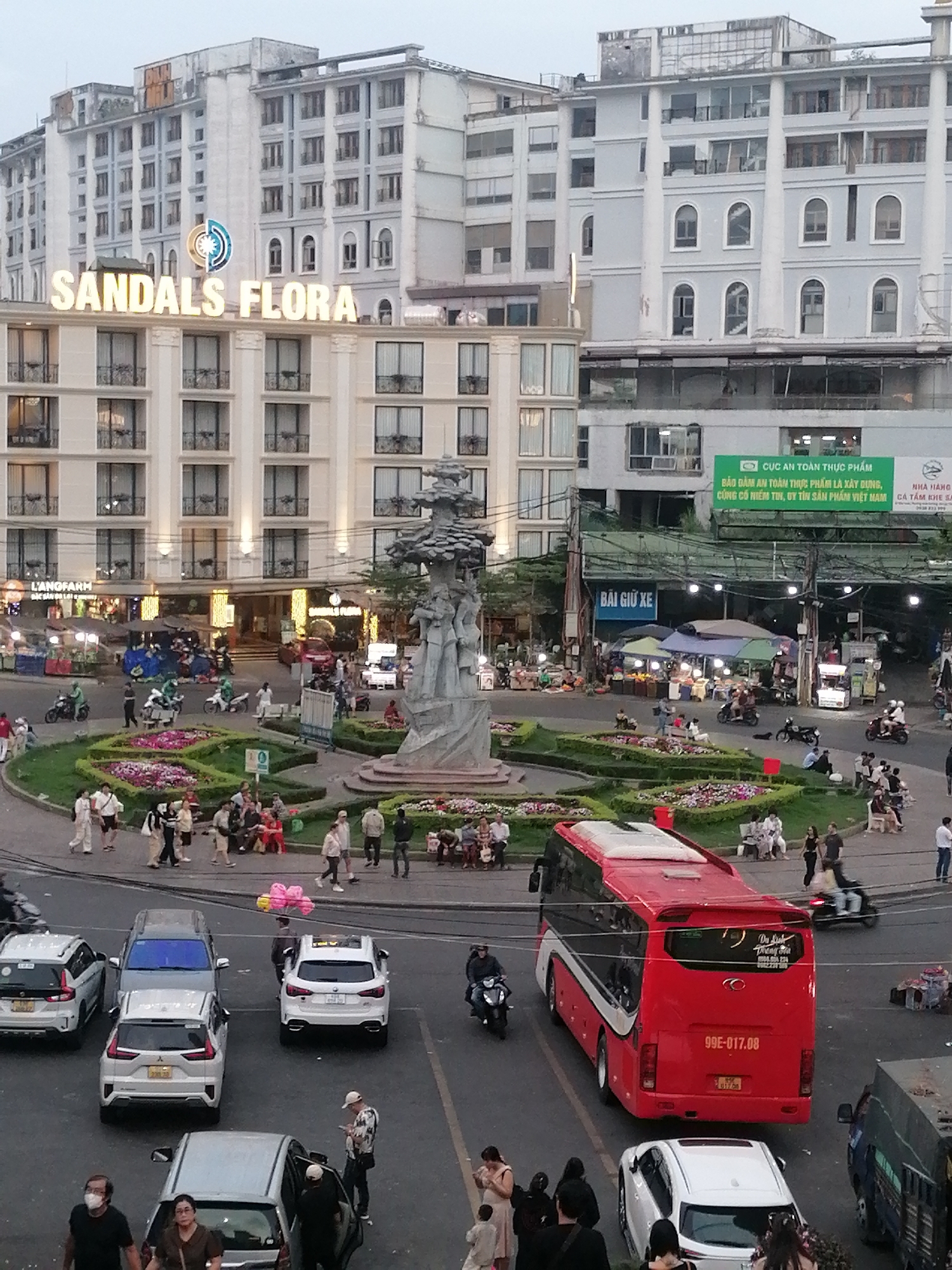 The image is a view of a busy roundabout in Da Lat, Vietnam. In the center of the roundabout is a statue surrounded by a flower garden where people are sitting and walking. The "Sandals Flora" hotel is a prominent building in the background. Other buildings, including one with a large green sign related to food safety, are also visible. A red bus is located in the foreground, along with several cars and motorbikes navigating the roundabout. Pedestrians are crossing streets, some wearing masks. Vendors and stalls are also present, indicating commercial activity in the area.