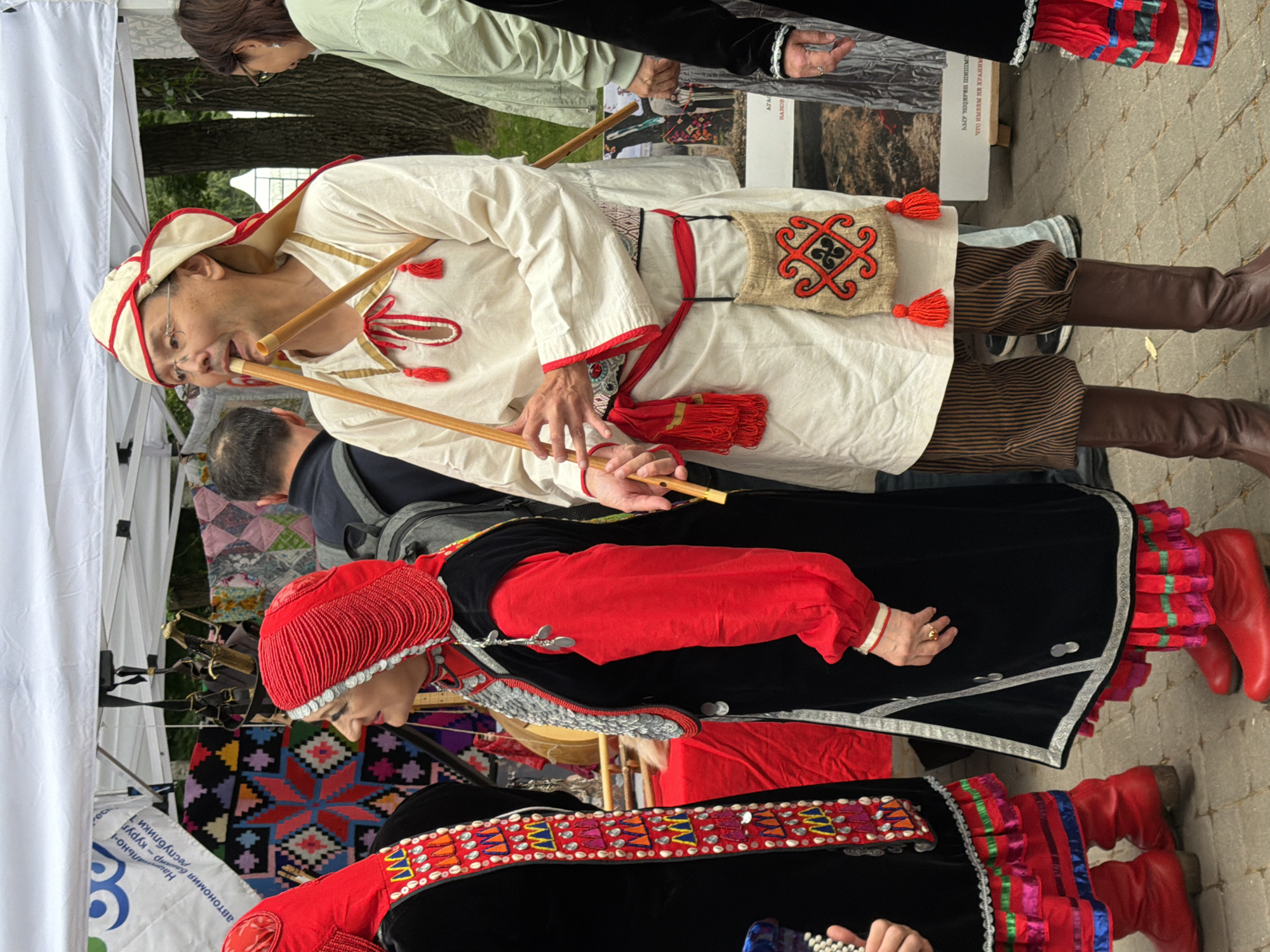 In Kazan, Russia, at an outdoor cultural event, two individuals are the primary focus. To the left, a woman in a traditional black and red outfit with an elaborate beaded headdress and silver adornments stands looking to the side. Her outfit features a black velvet overdress with embroidered trim and a bright red undershirt with puffed sleeves. Red leather boots complete her ensemble. To her right, a man in a white tunic and a distinctive white and red headdress plays two wooden flutes. He wears striped brown trousers and brown leather boots. A small, woven pouch with red and black embroidery hangs from his waist. The background reveals tents, colorful textile displays, and other attendees, suggesting a festival or market setting. The ground is paved with cobblestones, and trees are visible in the distance.
