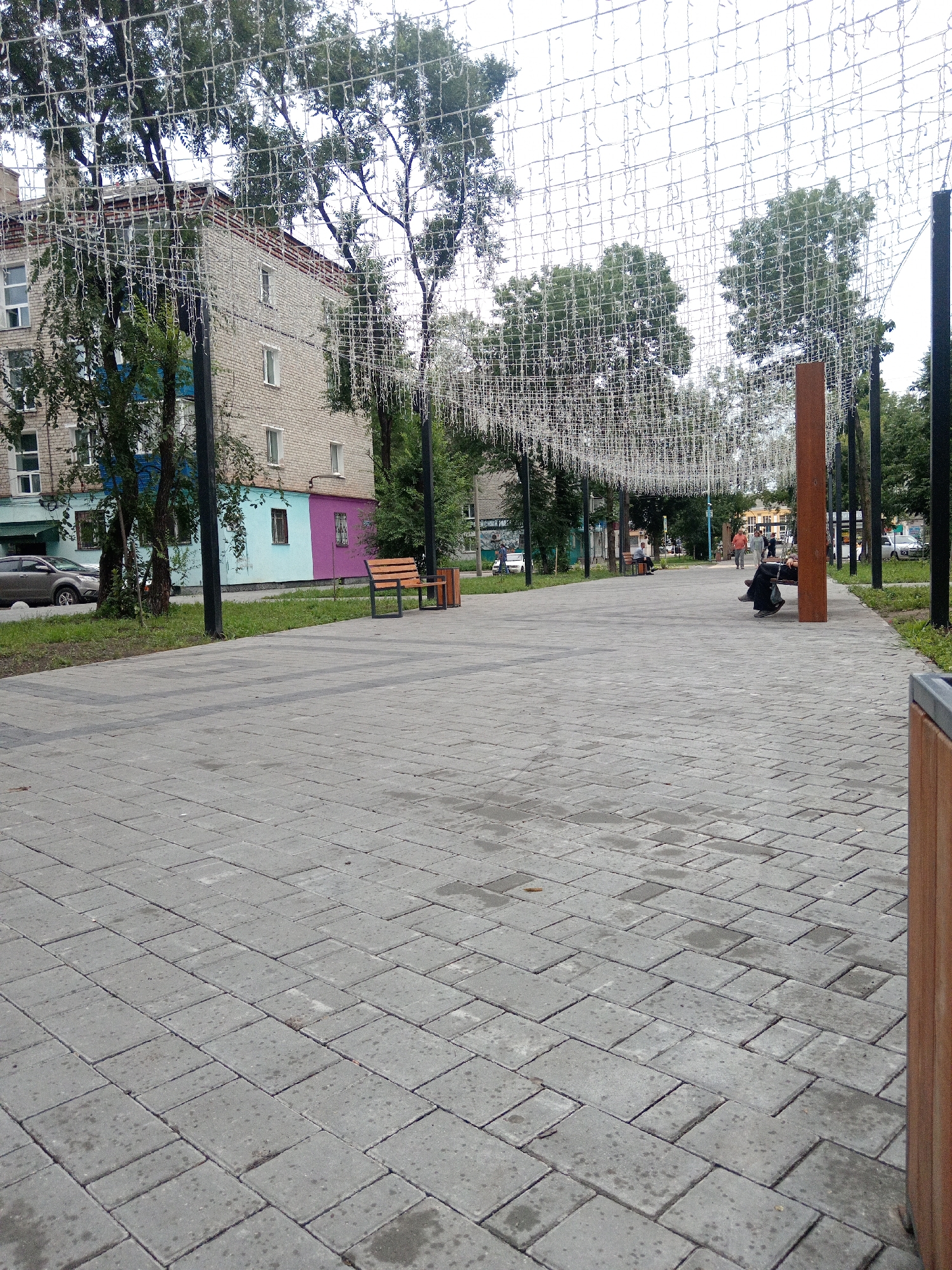 The image depicts a paved walkway in a city park, likely in Belogorsk, Russia, due to the distinctive building architecture and overhead string lights, which are common in Russian urban parks. The walkway is constructed of grey cobblestones laid in a patterned fashion. On the left side of the walkway, a multi-story building is visible, painted in segments of blue, teal, and purple. Trees line the building, and a car is parked on the street in front of it.
Further down the walkway, on the right side, there is a bench and a trash receptacle. A large, vertical wooden post stands near the middle of the walkway, serving as a visual marker. In the distance, several people are walking and sitting on benches, suggesting a public recreational area. The sky is overcast, and the overall lighting indicates it is daytime. The overhead string lights, draped across the walkway, are a prominent feature, possibly for decorative or celebratory purposes. The environment appears to be an urban park or pedestrian zone designed for public use.