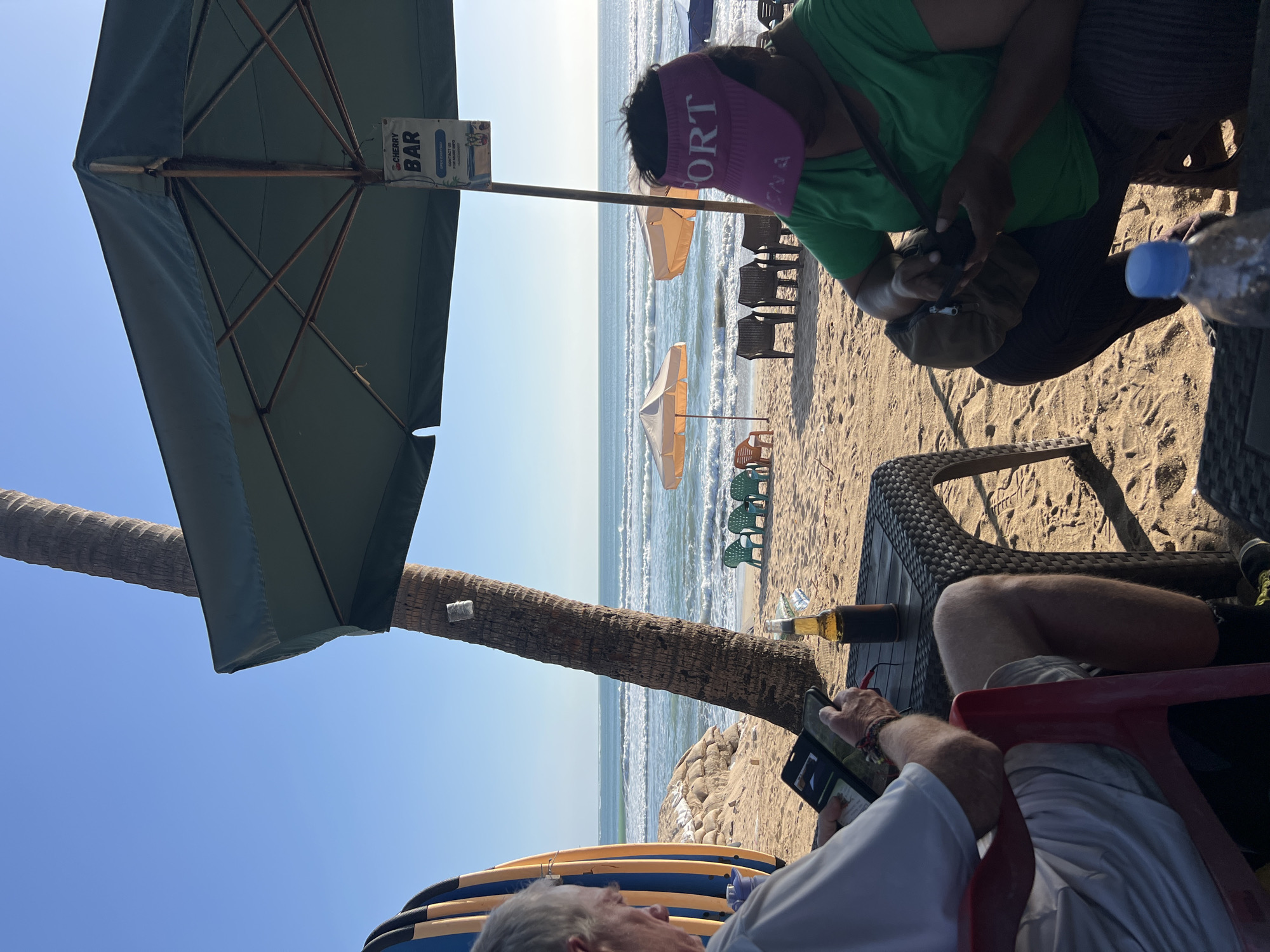An overhead shot captures a serene beach scene in Kuta, Indonesia, under a clear blue sky. A lone palm tree stands tall, its rough bark contrasting with the smooth texture of the sand. Beneath a large, dark green umbrella, two individuals are engrossed in their activities. On the left, an older man, wearing a white t-shirt and shorts, sits in a red chair, his attention focused on a tablet. A bottle of beer rests on a dark wicker table in front of him. To his right, a person in a green shirt and a pink visor, with "SPORT" emblazoned on it, crouches down, examining the contents of a dark bag. The beach stretches out before them, dotted with rows of empty plastic chairs and orange and beige beach umbrellas, suggesting a quiet moment before or after a busy period. The ocean, with its gentle waves, forms a tranquil backdrop to the scene, its surface shimmering under the sun. The overall atmosphere is one of relaxed leisure, a perfect representation of a day spent at the beach.