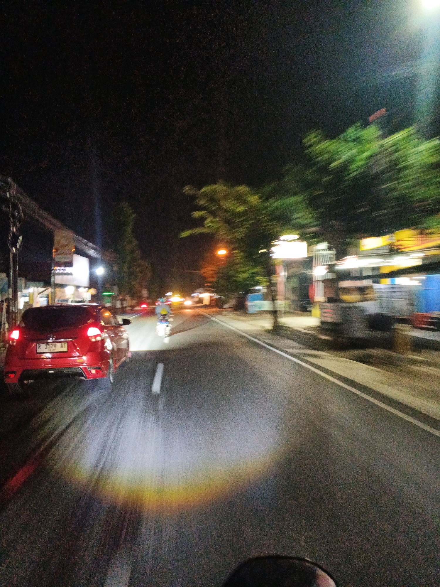 The image captures a motion-blurred night scene on a city street in Kroya, Indonesia, as indicated by the license plate "R 7579 AT" which belongs to the Banyumas region, where Kroya is located. The perspective suggests the photo was taken from a moving vehicle, likely a motorcycle, looking forward. A red car is prominently visible in the foreground, its taillights illuminated and its license plate legible. The car's headlights cast a bright beam onto the asphalt, creating a colorful, rainbow-like effect on the road surface, a phenomenon likely caused by the interaction of light with water droplets or exhaust.
The street is lined with buildings and trees, most of which are blurred due to the motion. Some of these buildings appear to be commercial establishments, with illuminated signs and storefronts visible. Traffic is present, with a motorcycle rider in the middle ground also illuminated by vehicle lights. The overall atmosphere is that of a typical urban night scene with moderate traffic and illuminated infrastructure. The sky is dark, indicative of nighttime, with a bright light source, possibly a street lamp or the moon, visible in the upper right corner. The image conveys a sense of movement and the ephemeral quality of light at night in an urban environment.