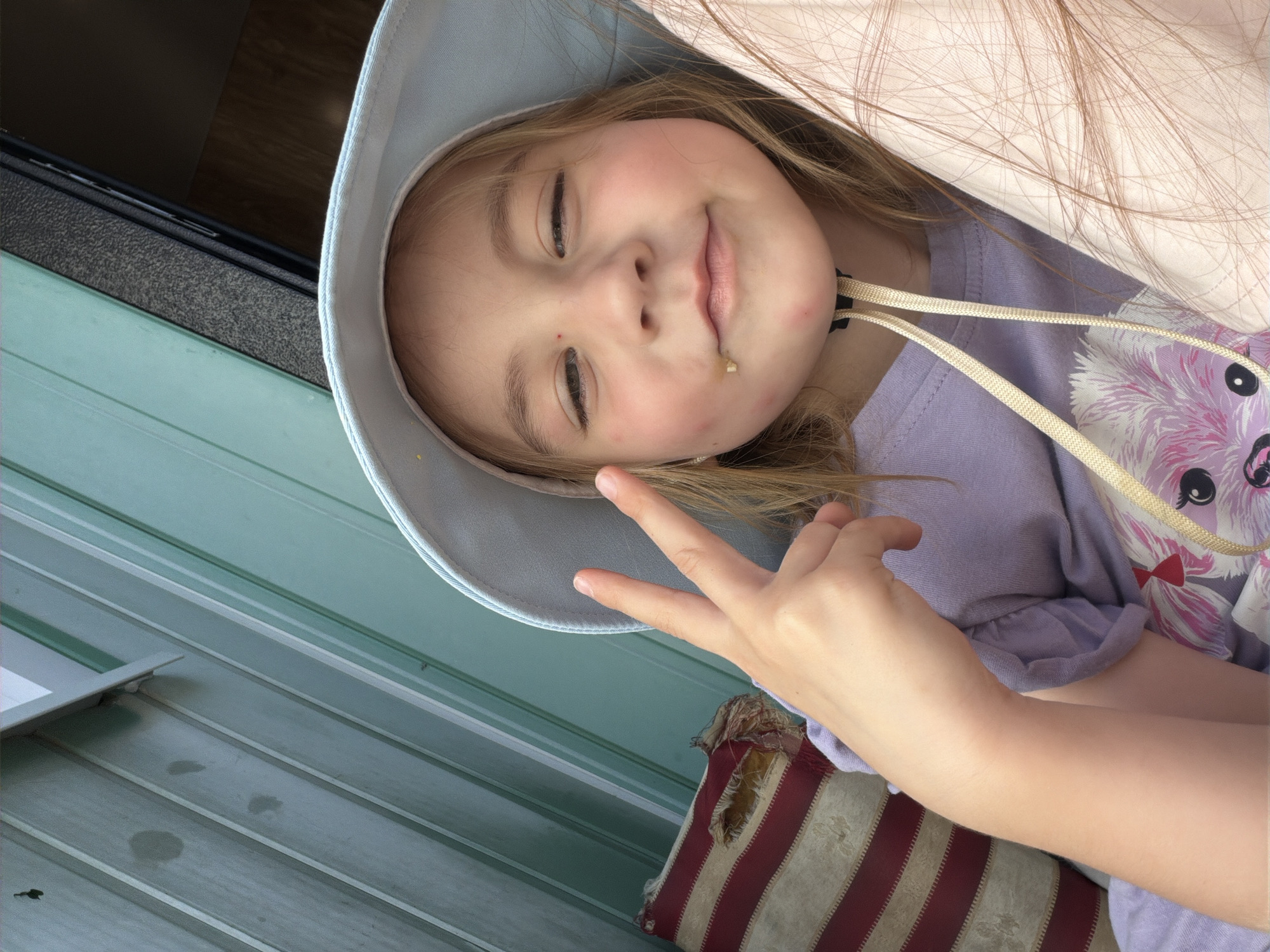 A young girl wearing a light blue sun hat and a purple t-shirt is captured in a close-up, candid shot. She is smiling with her eyes closed, a piece of food visible on her chin. Her right hand is raised, forming a peace sign. The girl's blonde hair is slightly visible around her face and falling over her shoulder. She is leaning against a light pink pillow or fabric. In the background, a teal-colored ribbed wall or siding is visible, suggesting an outdoor setting like a porch or patio. The lighting appears to be natural, possibly indicating daytime. The overall mood is cheerful and playful.
