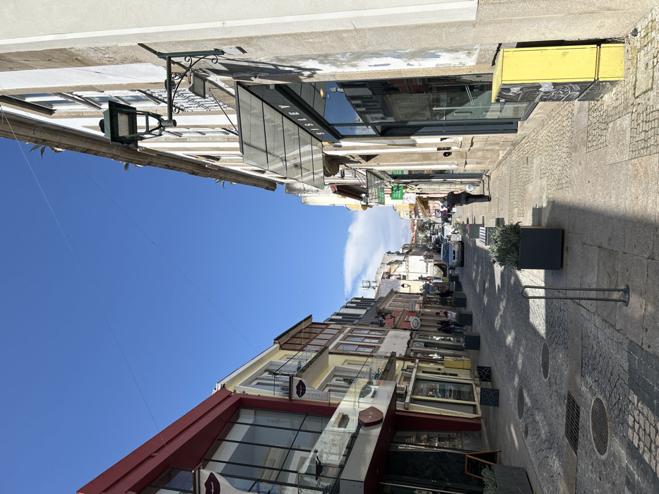 The image depicts a narrow urban street in Chaves, Portugal, viewed looking down its length under a clear blue sky. The street is lined on both sides by multi-story buildings with commercial establishments on their ground floors.
On the left, the foreground building features a prominent red upper facade with large reflective glass windows. Further down, buildings exhibit varying architectural styles and colors, including yellow, light brown, and white, with some possessing balconies. Their ground levels contain multiple storefronts.
On the right, a light-colored building with vertical stone-like stripes dominates the foreground. It features a large glass storefront with a transparent awning displaying "SISLEY." An ornate green street lamp is affixed to this building. A bright yellow utility box is visible at the building's base. Further down, other storefronts are present, including one with a green sign.
The street itself is paved with cobblestones, featuring several circular manhole covers and rectangular grates. Concrete sidewalks border both sides of the street. Dark grey rectangular planters containing green foliage are spaced along both sidewalks, some accompanied by benches. Metal bollards are also present on the sidewalks.
One individual, dressed in dark attire, is visible walking away from the camera on the right sidewalk. Several other indistinct figures appear as pedestrians further down the street on the left. Multiple passenger vehicles, including a light-colored sedan, are visible parked or in motion further down the street.
Overhead wires traverse the sky, which is a clear blue with a small patch of white cloud visible in the distance. Shadows from the buildings extend across the street and sidewalks, indicating bright daylight conditions. A communication tower structure is faintly visible on a distant rooftop.