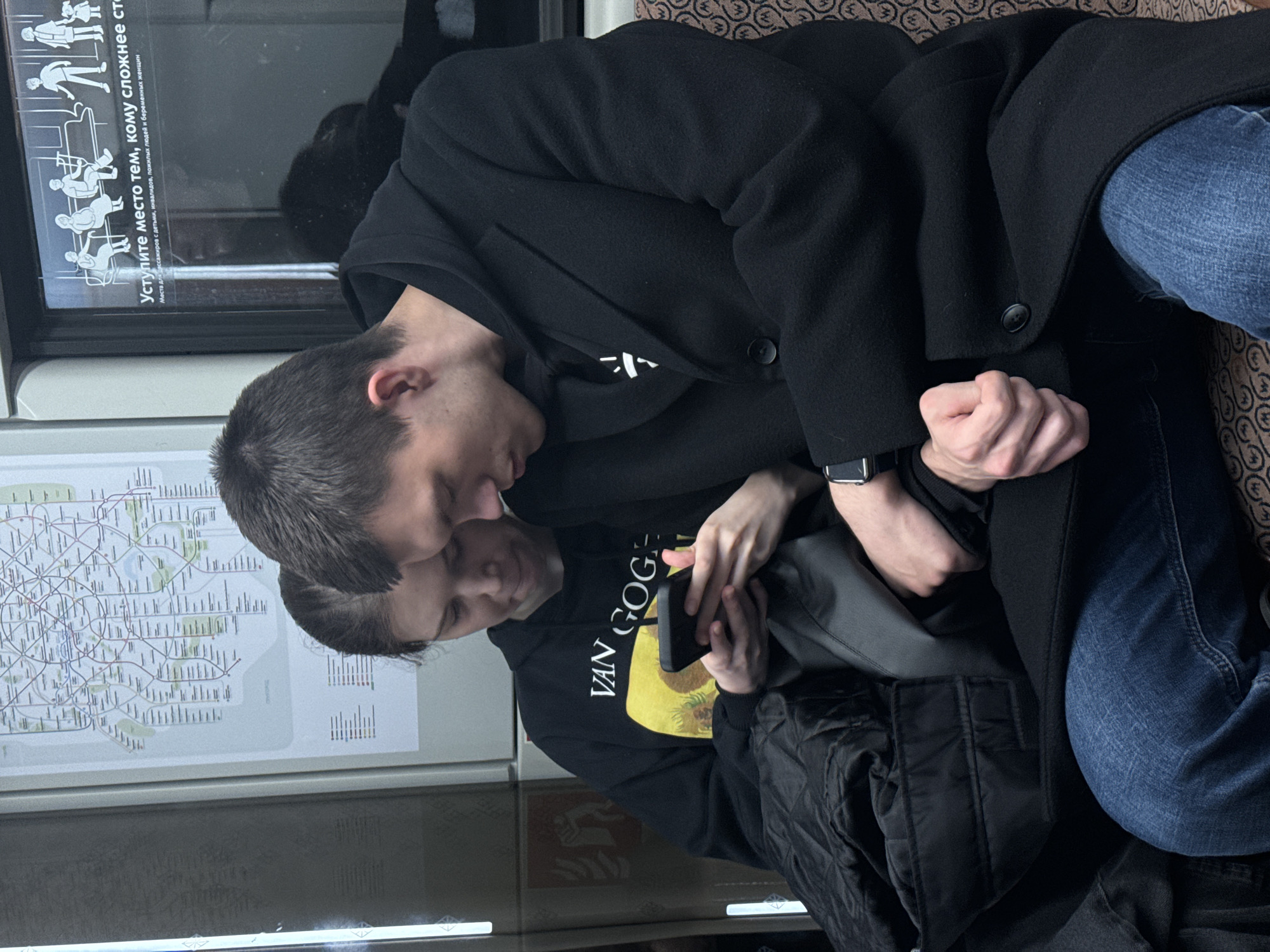 Two men are sitting next to each other on what appears to be a train. The man on top is wearing a black coat and jeans, with a watch visible on his wrist. The man below is wearing a black hoodie with "VAN GOGE" printed on it, along with a yellow image. He is holding a mobile phone. The train appears to be in Moscow, Russia, as indicated by the presence of a Moscow metro map. The sign above reads “Уступите место тем, кому сложнее ст” meaning “Give way to those who have more difficulty st”, with the rest of the sentence cut off.