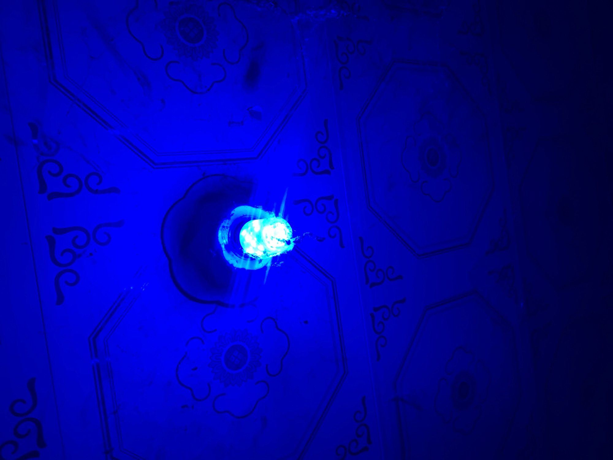 A low-angle shot shows a blue-lit ceiling, perhaps in the New Stadium area of Nigeria. The ceiling has a pattern of connected hexagons, each containing a stylized floral design and small curved flourishes on the edges. The hexagons are delineated by darker blue lines. A bright, roughly circular light source sits in the center-left of the frame, casting blue light on the ceiling. The right side of the image is significantly darker than the left.