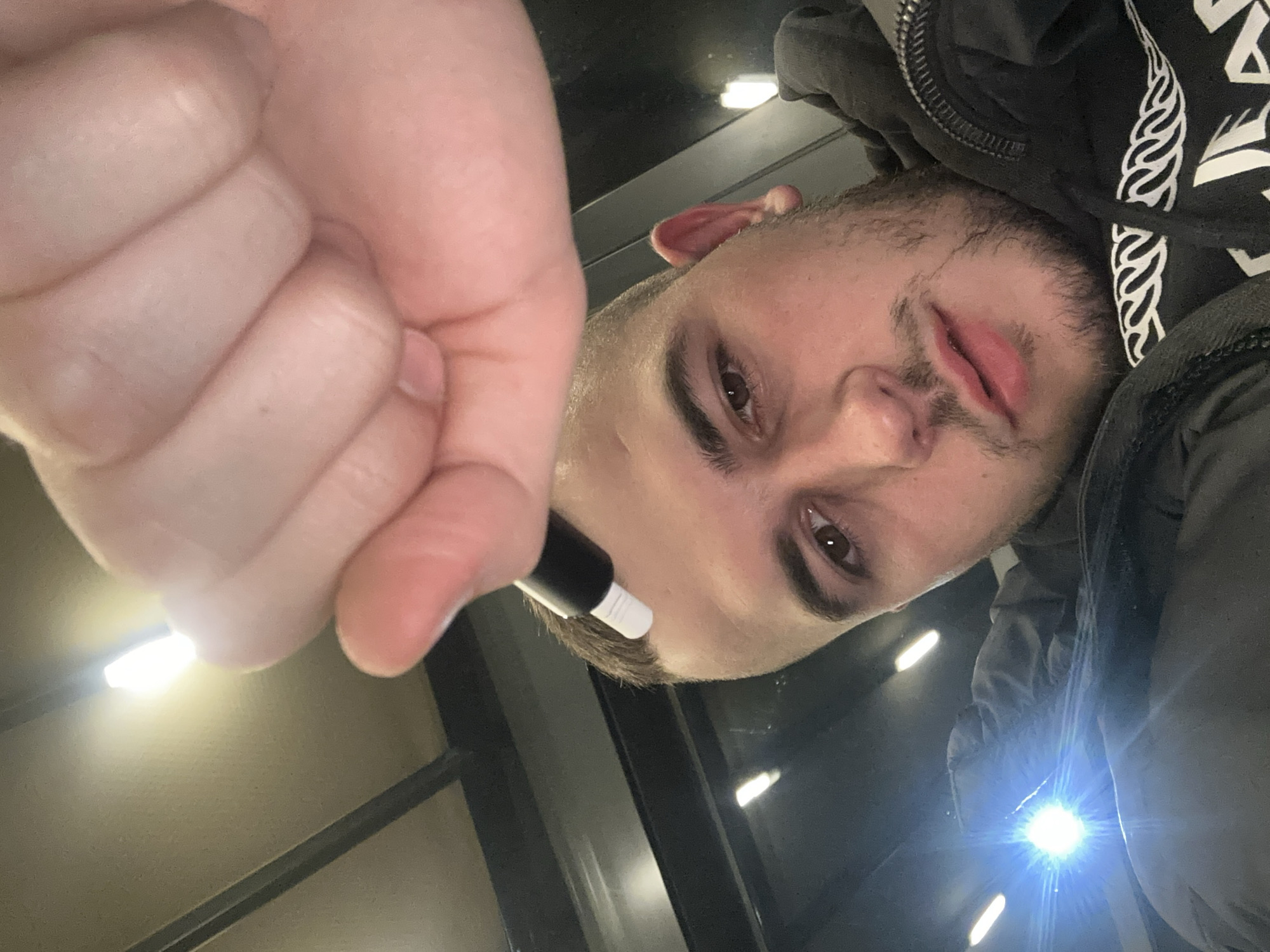 A selfie taken from a low angle shows a light-skinned male holding a black and white pen near his face. The man has short brown hair and a beard. He is wearing a black hooded jacket with white designs. The background shows a ceiling with rectangular lights. The location appears to be indoors and there are no features that could pinpoint the city of Plovdiv.
