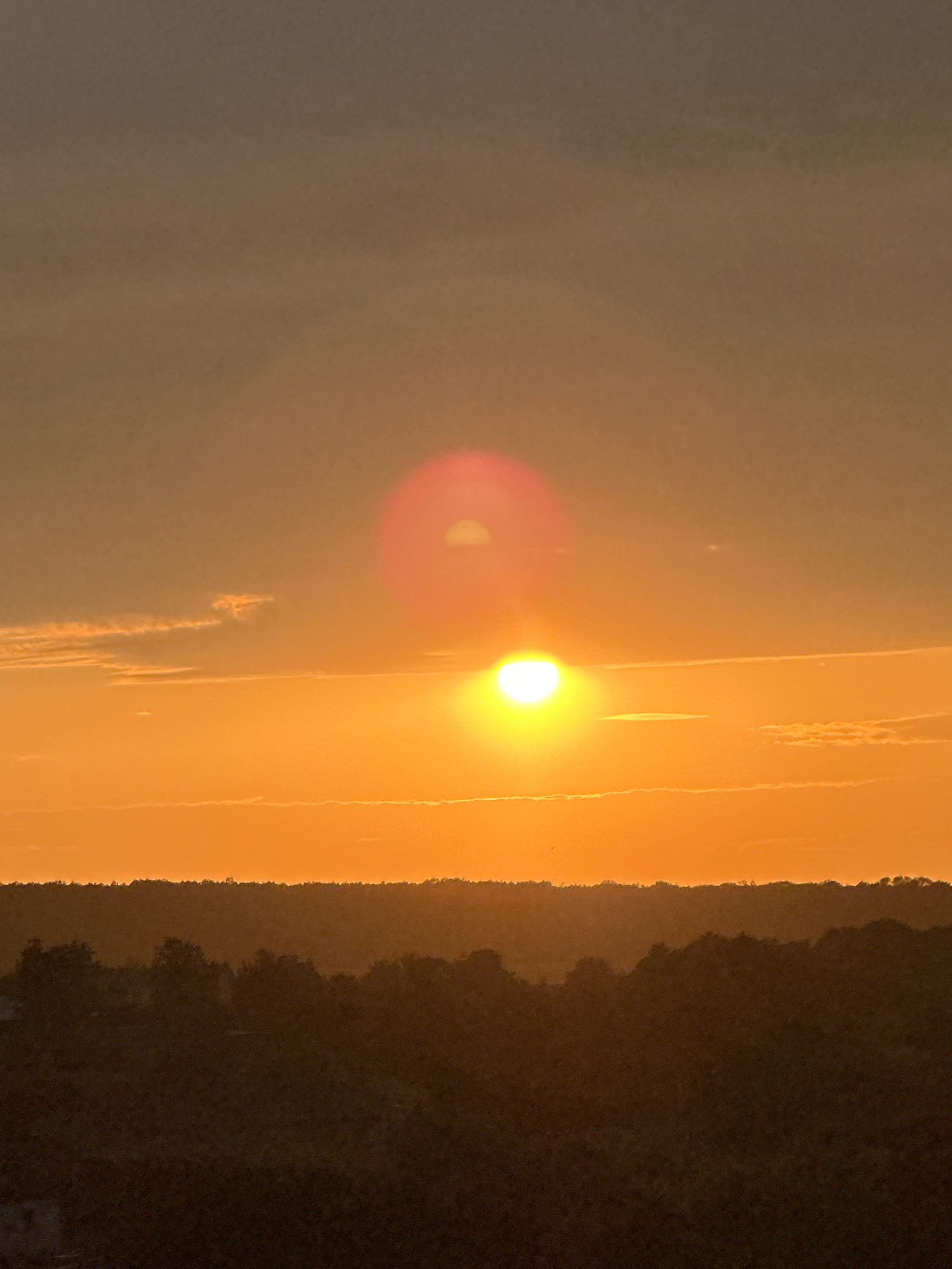 This is a photograph of a sunset over a rural landscape. The sun is a bright, glowing orb in the center of the frame, casting a warm orange and yellow light across the sky. There are some wispy clouds in the sky, tinged with the colors of the sunset. Below the sky, there is a dark silhouette of trees and rolling hills, suggesting a natural, undeveloped area. The overall impression is one of a peaceful and serene evening. There is no indication of the city of Makiv, Ukraine, or any specific urban features.