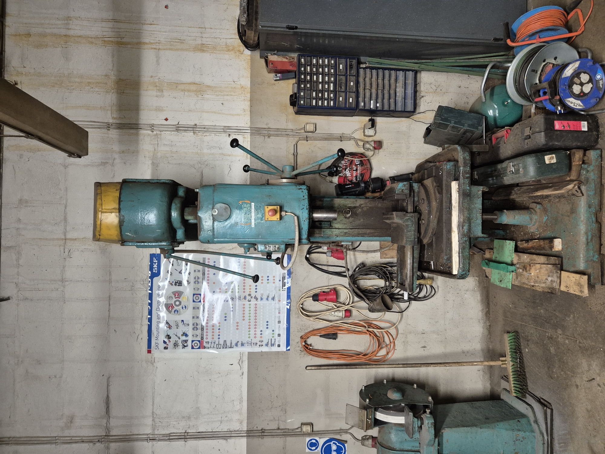 The image depicts a workshop environment in Ebavere küla, Estonia. A large, vintage turquoise and yellow drilling machine dominates the center of the frame. It has a yellow cover on top, multiple handles, a red emergency stop button, and a metal vise assembly. To the left of the drilling machine, a smaller turquoise grinding machine is visible. The walls are concrete, with some staining and visible conduits. Hanging on the wall above the drilling machine is an SKF poster detailing mounting and dismounting procedures. Several electrical extension cords are coiled and hung on the wall, along with a broom and some safety signs with eye and ear protection symbols. To the right of the drilling machine, a blue multi-drawer cabinet filled with small items is stacked on top of another, more transparent drawer unit. A tall, dark grey cabinet stands to the far right. On the floor near the drilling machine, there are various items including a green toolbox, several wooden planks, a green air tank, a coiled orange extension cord reel, and a blue and grey electric cable reel. The floor is concrete and appears dusty and somewhat stained, consistent with a working workshop. There are no people visible in the image.