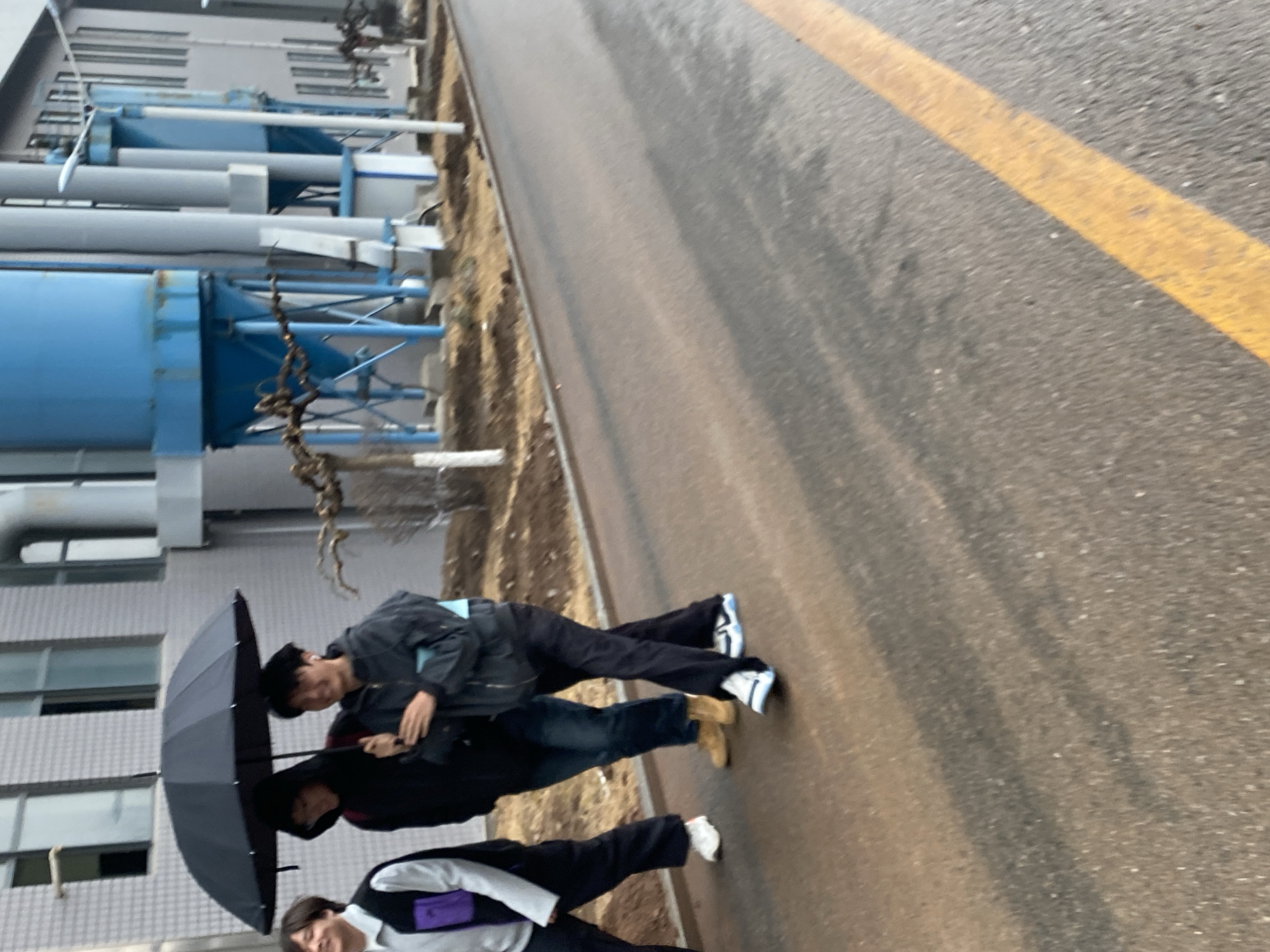 The image depicts a street scene in Zhongtun, China. A building with multiple windows and what appears to be industrial equipment (likely water tanks and pipes) stands to the left. Three people are walking along the right side of the street under an umbrella. The pavement is wet, and there's a yellow line marking the edge of the road.