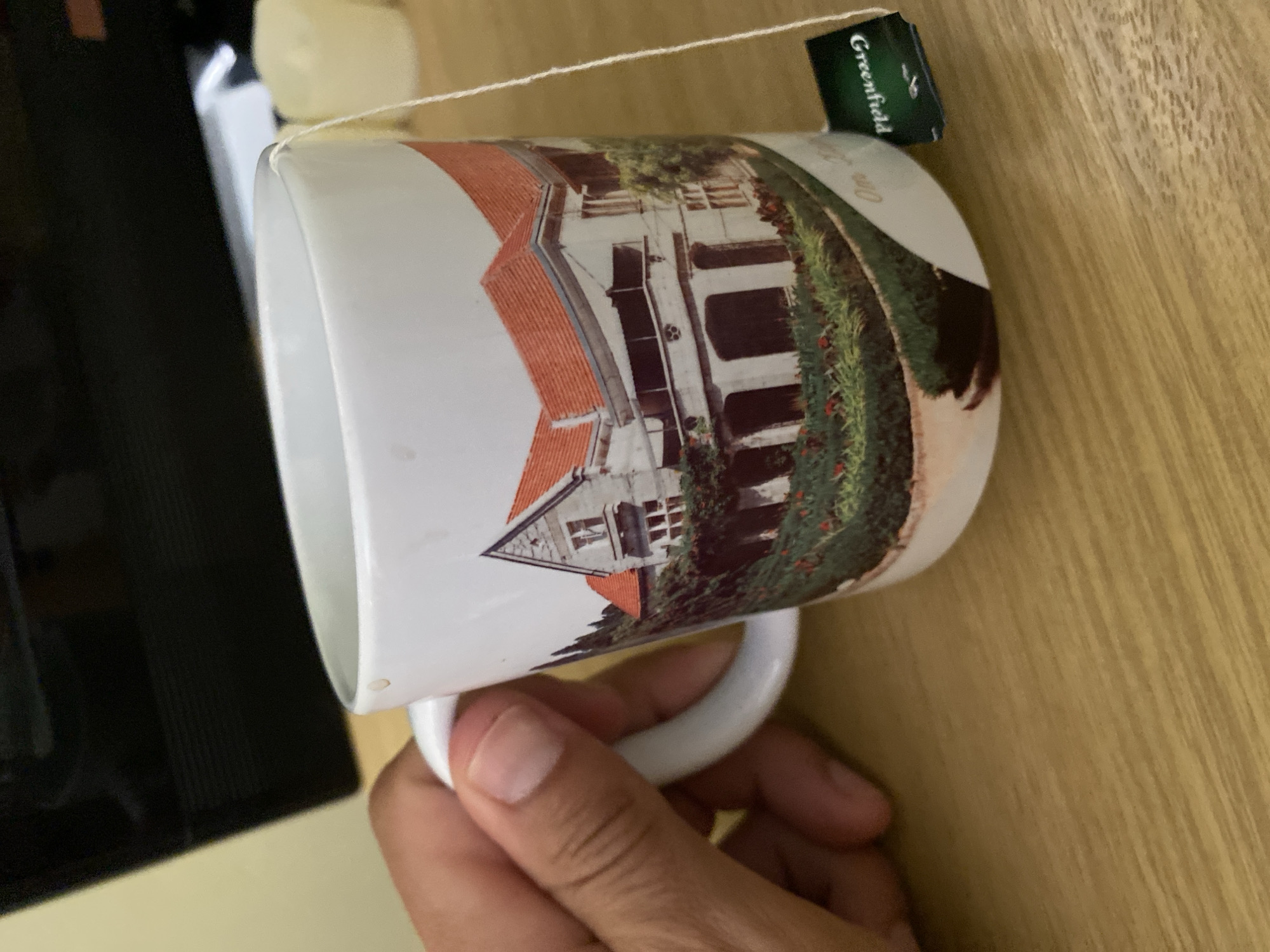 A hand is holding a white ceramic mug. The mug features a printed image of a large house with a red-tiled roof, surrounded by greenery and flowers. A tea bag string hangs over the edge of the mug, with a green tea tag labeled "Greenfield" visible. The mug sits on a wooden surface, likely a desk or table. In the background, a dark rectangular object, possibly a monitor, is visible, along with two out-of-focus cylindrical objects, likely candles. The scene appears to be indoors, with soft lighting. There is no indication of the specific location within Гаспринский поселковый совет, Ukraine.