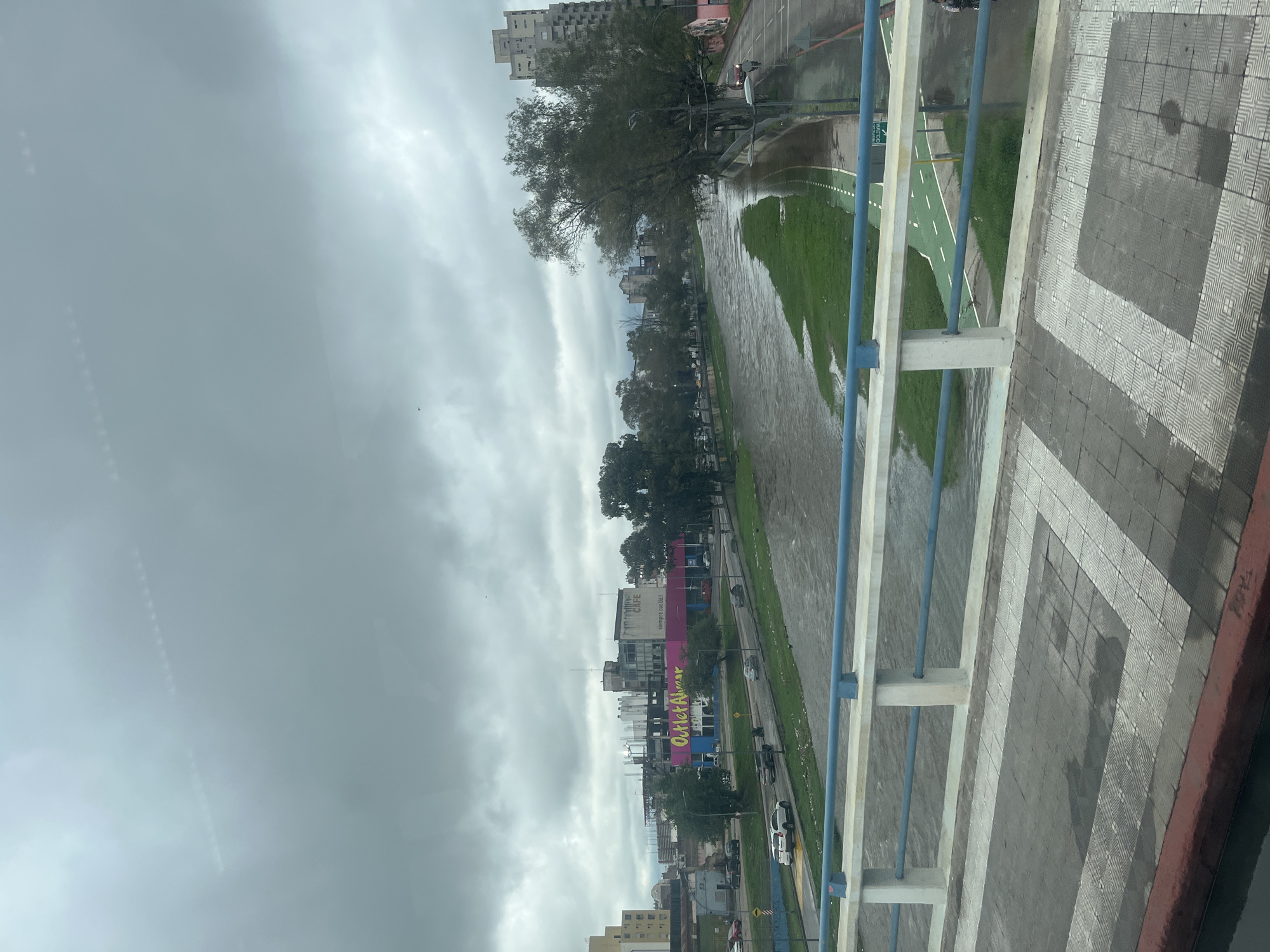 The image shows an overcast day in Córdoba, Argentina, with a view from a bridge overlooking a river. The river appears swollen, possibly due to recent rainfall. A paved path with white and green markings runs alongside the riverbank. On the opposite side of the river, buildings line the street, including a large building with the words "Outlet Almacen" painted in large, bright letters. Traffic can be seen moving along the road. The bridge itself has blue railings and a patterned grey and white tiled walkway. No people are clearly visible in the scene.
