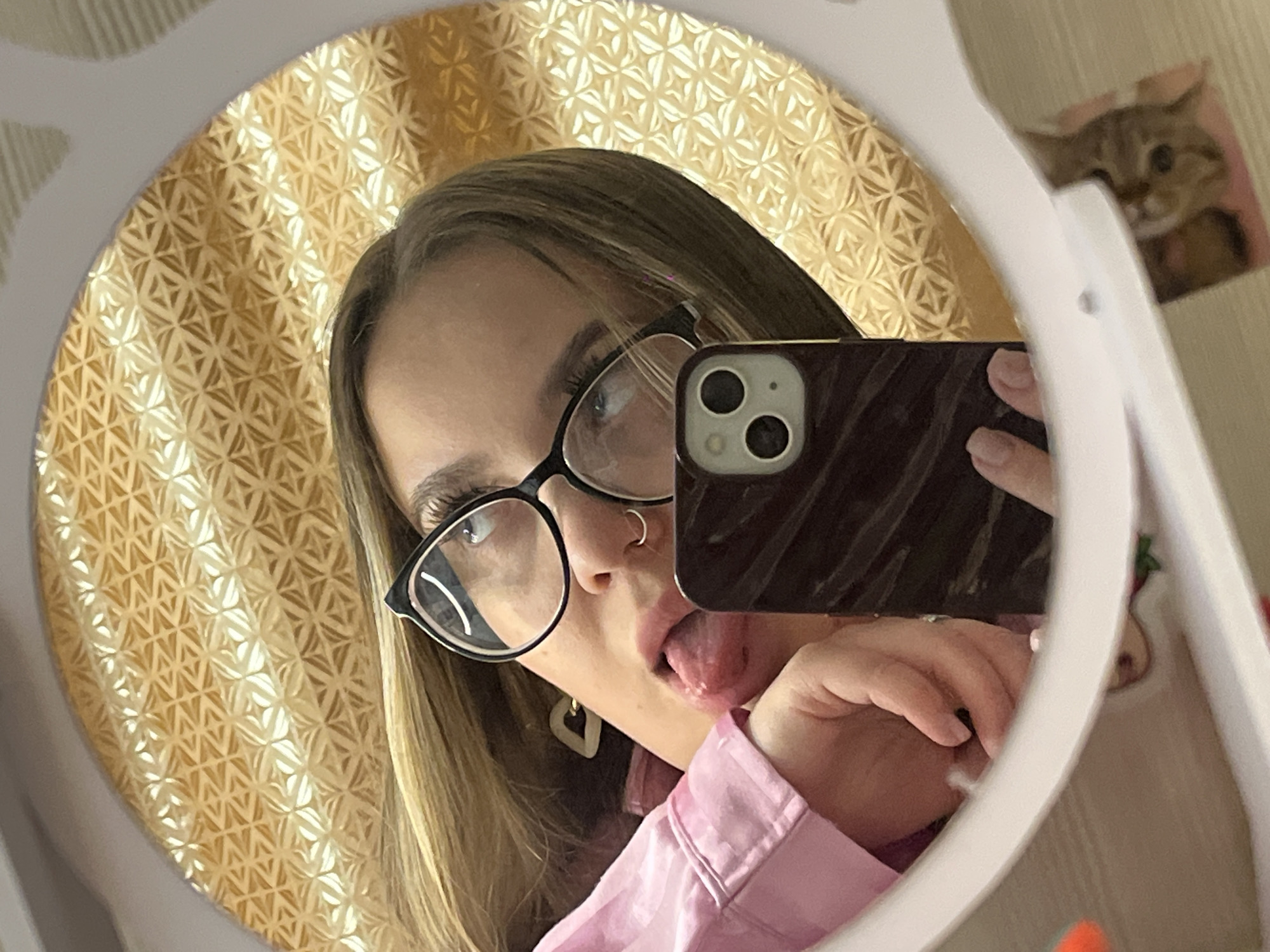 A woman is taking a selfie in a mirror. She is wearing glasses, a nose ring, and a pink shirt. She is sticking her tongue out. The background is a gold patterned wall. A cat picture is visible in the top right corner. The location is Novopokrovskaya, Russia.