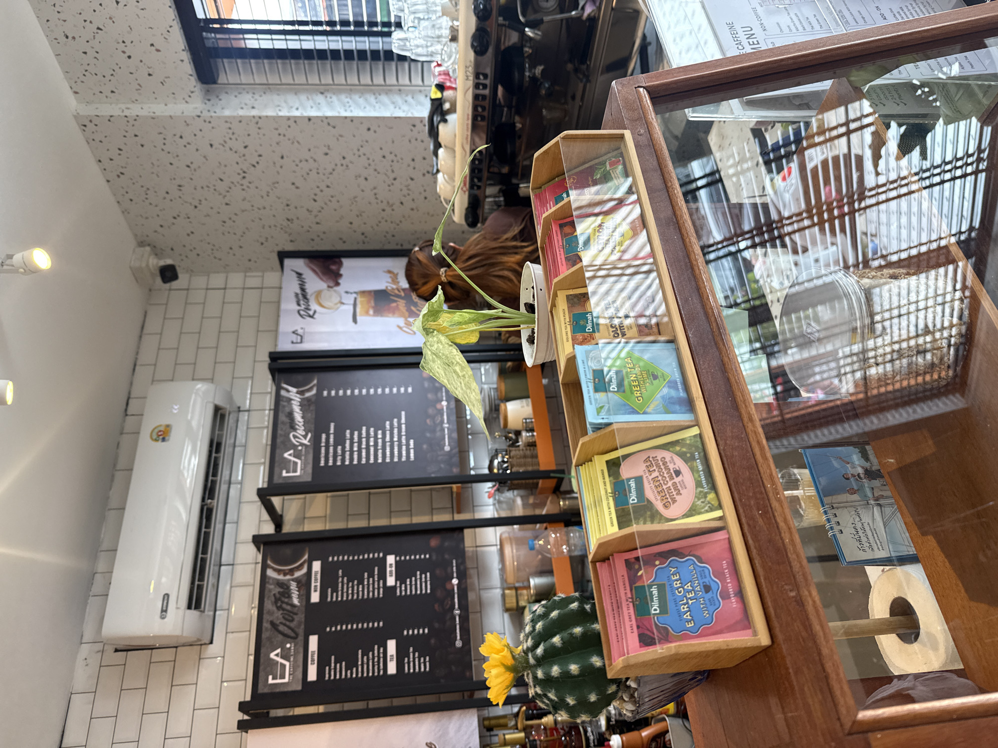The image depicts the interior of a cafe in the city of Ko Samui District, Thailand. In the foreground, a wooden display case holds various flavors of Dilmah tea bags, including Earl Grey with Vanilla, Green Tea with Coconut and Mango, and Green Tea with Lemon. To the left of the tea display, a cactus with a yellow flower is visible in a pot. Behind the tea display, a woman with long brown hair is seen from the back, facing a coffee machine. The walls are tiled in white brick and a speckled pattern. An air conditioning unit is mounted on the tiled wall. Two black menu boards are displayed on the wall; one lists coffee and non-coffee drinks, and the other appears to be a recommended menu. The overall environment suggests a well-lit and organized cafe setting.