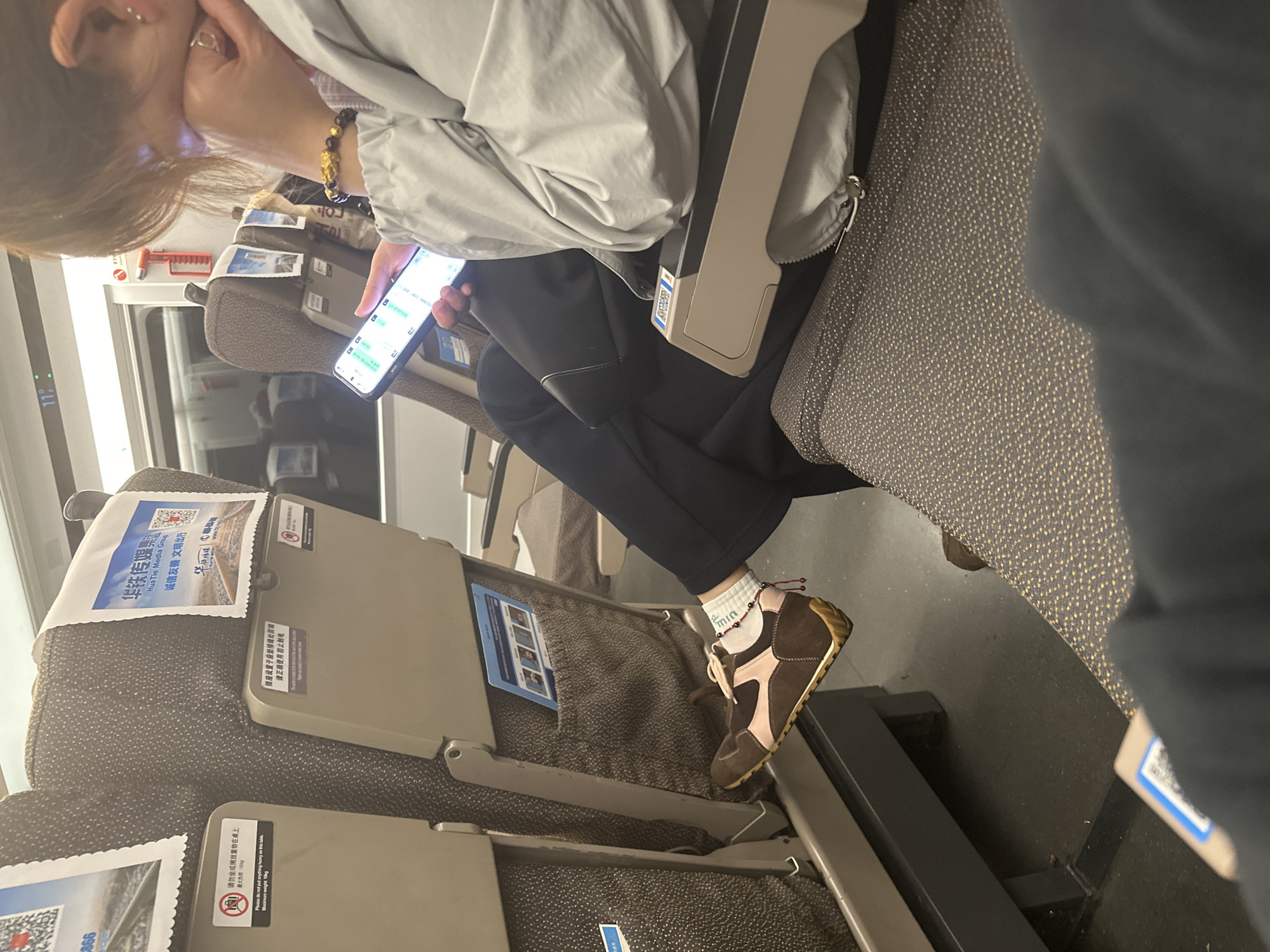 An indoor, eye-level shot captures a person seated on what appears to be a train or high-speed rail in Yuhang District, China, engrossed in their smartphone. The person has light brown hair and is wearing a light grey jacket, black pants, white socks, and distinctive brown and pink shoes. A gold bracelet adorns their right wrist, and a red beaded anklet is visible on their left ankle above the white sock, which features the text "min".
The individual is looking down at their phone, held in their hands, with their left hand positioned near their face. The phone's screen displays what looks like a messaging application or perhaps financial data with various numbers.
The setting is a public transport carriage with multiple rows of seats upholstered in a dark, textured fabric with small gold or light-colored flecks. Foldable tray tables are attached to the back of the seats. On the back of the seat in front, a white cover displays Chinese text for "Hua Tie Media Group" and "诚信友善文明出行" (Integrity, Friendliness, Civility, Travel) alongside QR codes. Another sticker warns, "请勿坐或搁放重物在桌上 最大负重 10kg" (Please do not sit or place heavy objects on the table, maximum weight 10kg). Another sign indicates, "插座设置于座垫接缝处前端 请正确使用防止触电" (Socket is located at the front of the seat cushion joint, please use correctly to prevent electric shock). A digital display on the wall shows "11", possibly indicating temperature or time. The lighting is artificial, suggesting an indoor environment, and the person appears relaxed and focused on their device.