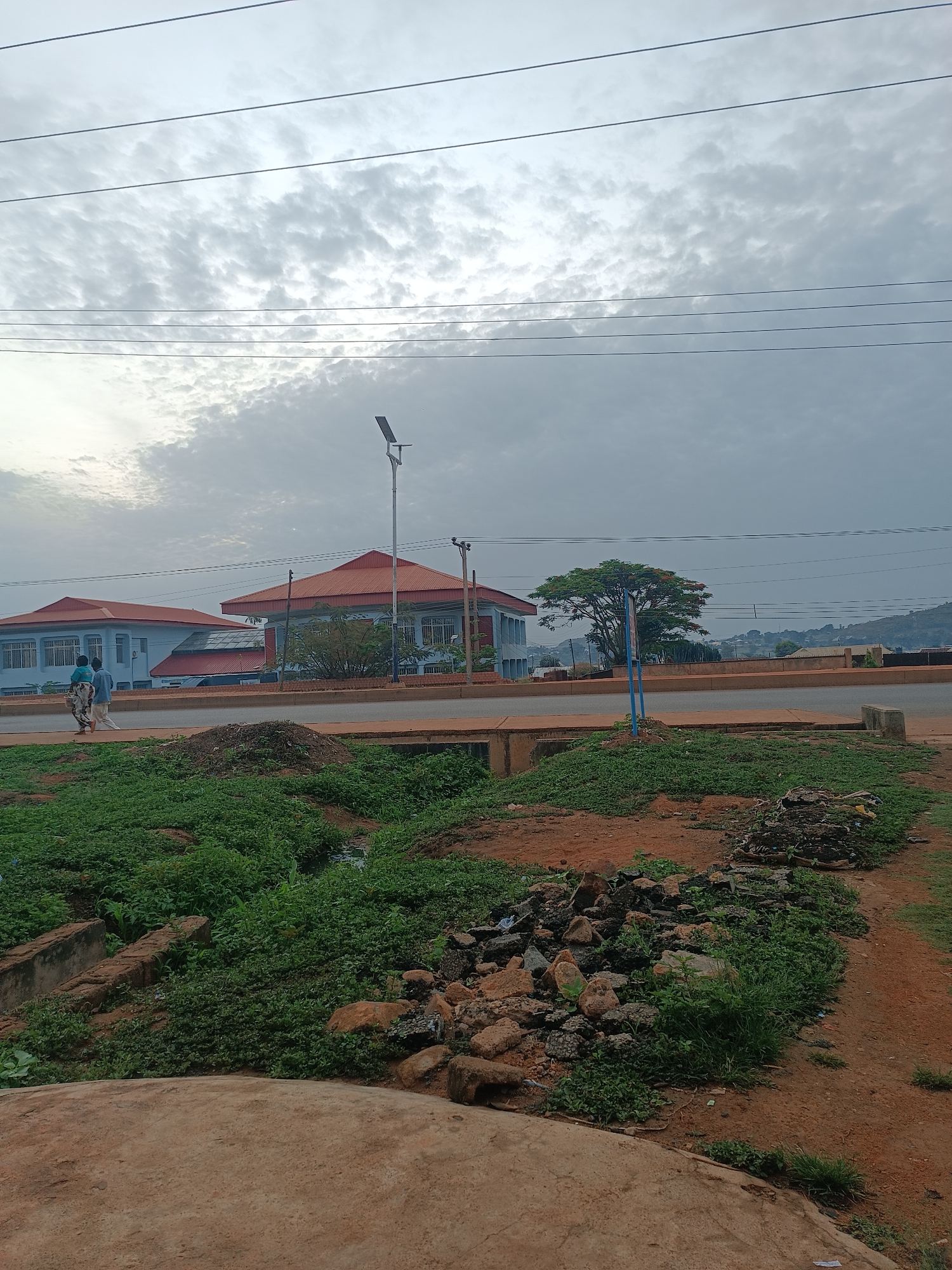 The image shows a street scene in Bukuru, Nigeria. Two light blue buildings with red-orange roofs are situated across a paved road. A solar street lamp stands tall in front of the buildings. Power lines stretch across the sky, which is overcast with dense clouds. In the foreground, a ditch filled with green vegetation and rocks runs alongside a red-brown dirt path. Two people are walking on the sidewalk across the street. Hills are visible in the distance.