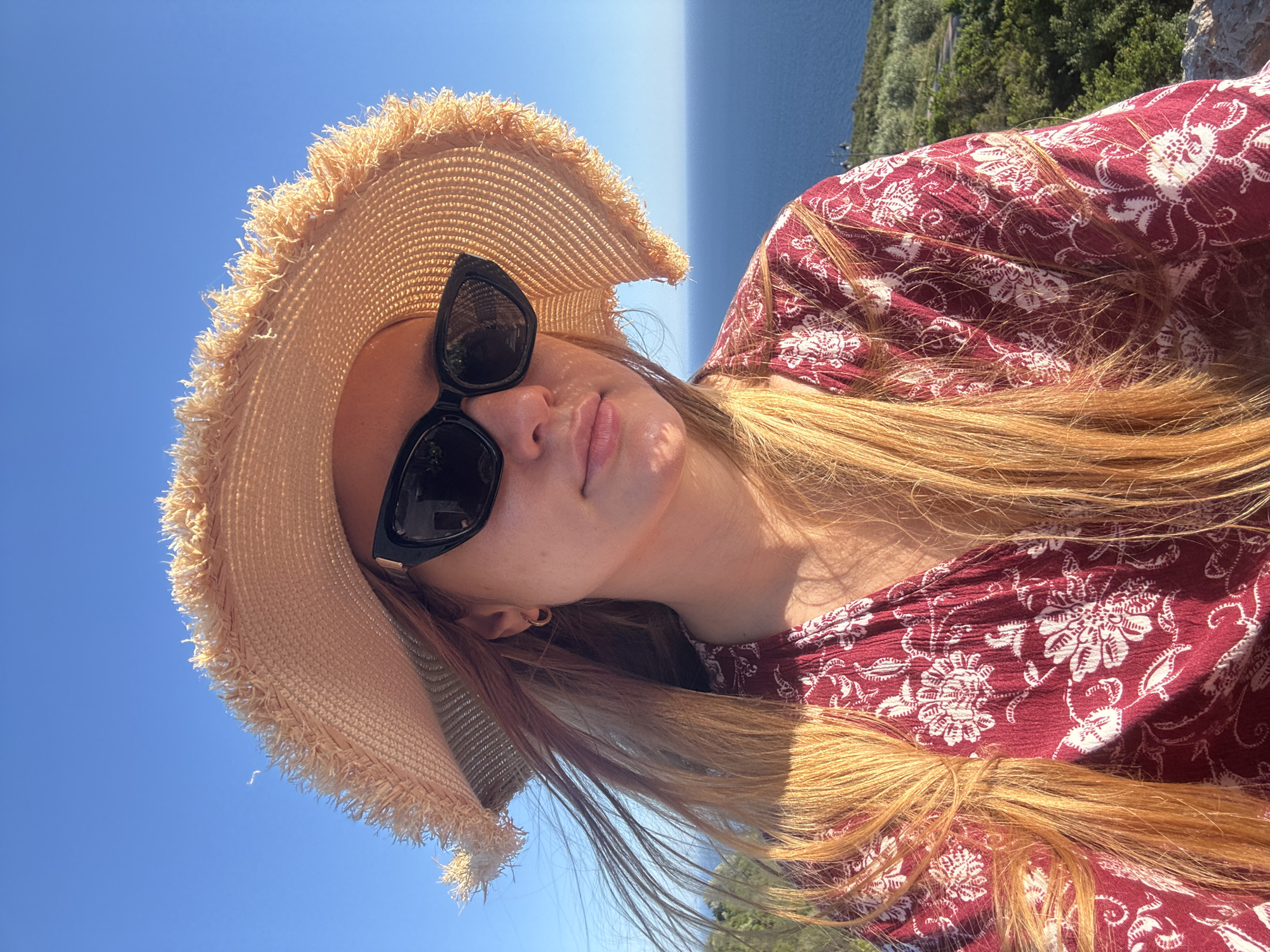 The image presents a head and shoulders shot of an adult female, illuminated by direct sunlight, indicating an outdoor setting in Krimovice, Montenegro. She wears a wide-brimmed, light tan straw hat with a frayed brim and dark, cat-eye style sunglasses. Long, light brown to ginger hair, highlighted by the sun, frames her face and extends over her left shoulder, with some strands appearing to be moved by a slight breeze. A small gold hoop earring is visible on her left earlobe. Her attire includes a dark red garment with a repeating pattern of white, intricate floral and paisley motifs. The background features a clear, deep blue sky dominating the upper portion, with a faint white streak visible near the hat. Below the sky line, a vast expanse of calm, deep blue sea extends towards the horizon on the right. Below the sea, the land reveals a lush green hillside covered with trees and shrubs. A paved road or path is visible winding through the greenery, and a street lamp is discernible on the far right, overlooking the landscape and sea.