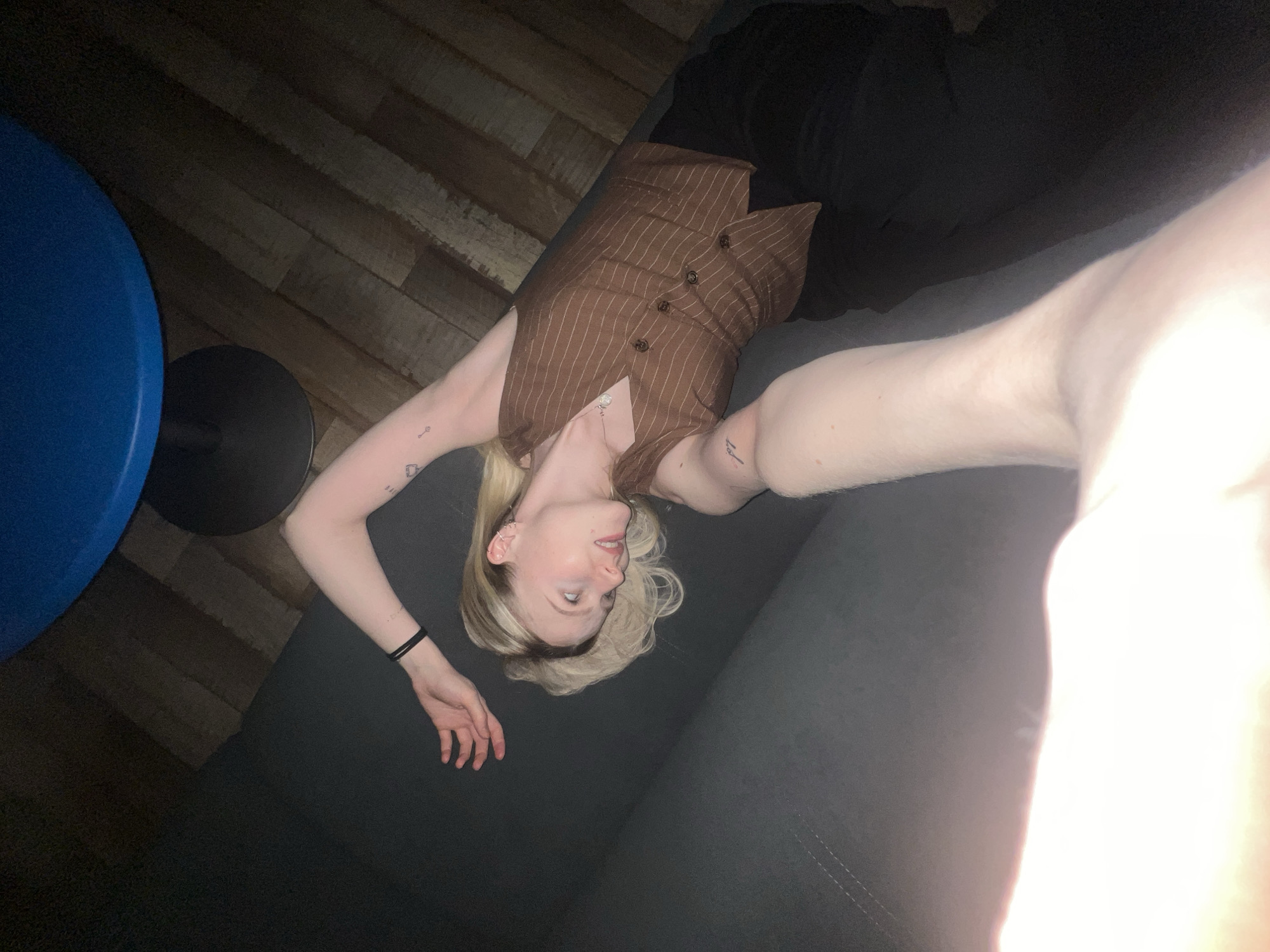 A person is lying on a dark gray couch in an indoor setting. They are wearing black pants and a brown pinstripe vest. The person has fair skin and blonde hair, with visible tattoos on their arms. A blue circular object with a black base is to the left. The background includes dark wood flooring. The location is not apparent from the image.