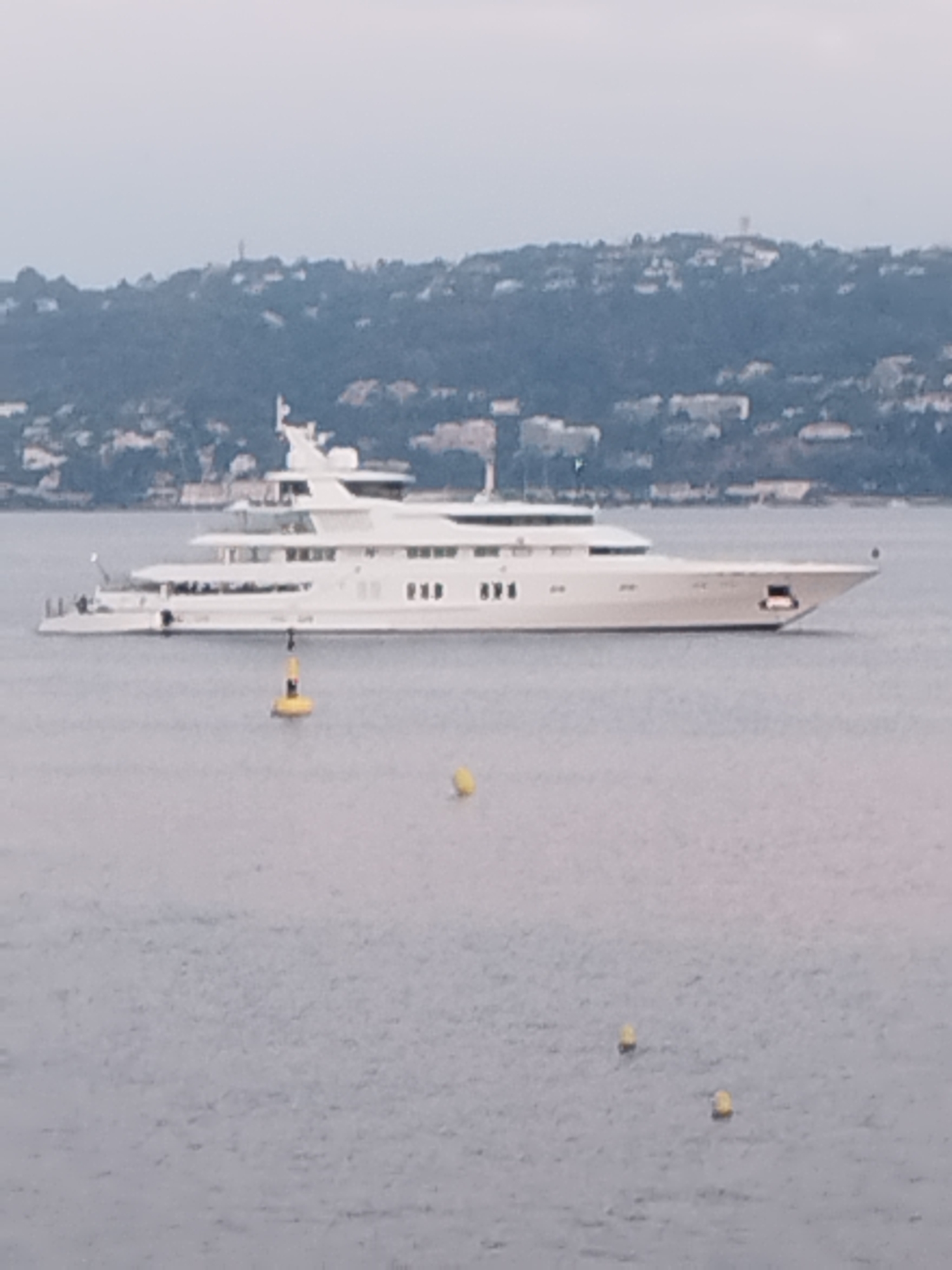 The image depicts a large, white, multi-deck yacht moored in the water. The yacht has a sleek, modern design with multiple windows and balconies. In the foreground, a yellow buoy with a red top floats in the water, attached to the yacht by a rope. In the background, a densely populated, tree-covered hillside is visible, dotted with buildings, suggesting a coastal urban or semi-urban environment. The sky is overcast and a light haze is present, creating a muted color palette. The scene is likely Antibes, France, given the characteristic Mediterranean coastline and the presence of such large yachts in its waters. There are no people clearly visible on the yacht or in the surrounding environment.