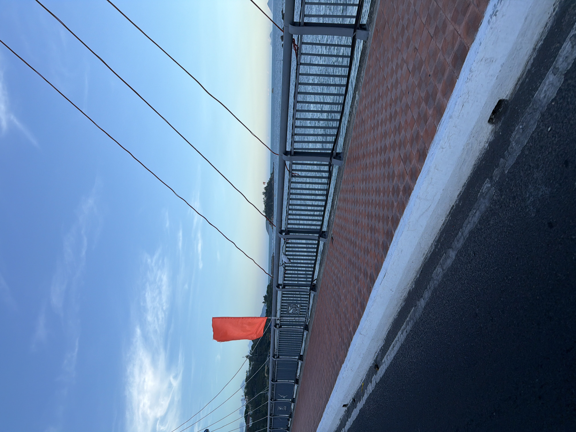 The image shows a bridge with metal railings and a brick path, positioned over water, possibly the coast. A bright orange flag is visible, seemingly attached to the bridge's structure. The scene is partially obscured by an asphalt road with a white curb in the foreground. Overhead electrical wires are also visible, stretching across the sky. The sky is light blue with scattered clouds. This is the city of Nha Trang Ward, Vietnam.