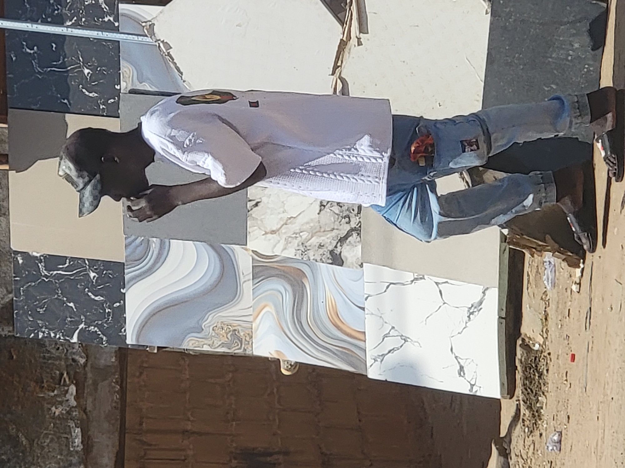 A man wearing a baseball cap, a white shirt, and blue jeans stands on a collection of square tiles in Maiburiji, Nigeria. The tiles have varying patterns and textures, including marbled designs in gray, white, black, and brown. The ground around the tiles appears to be a mix of dirt and debris.