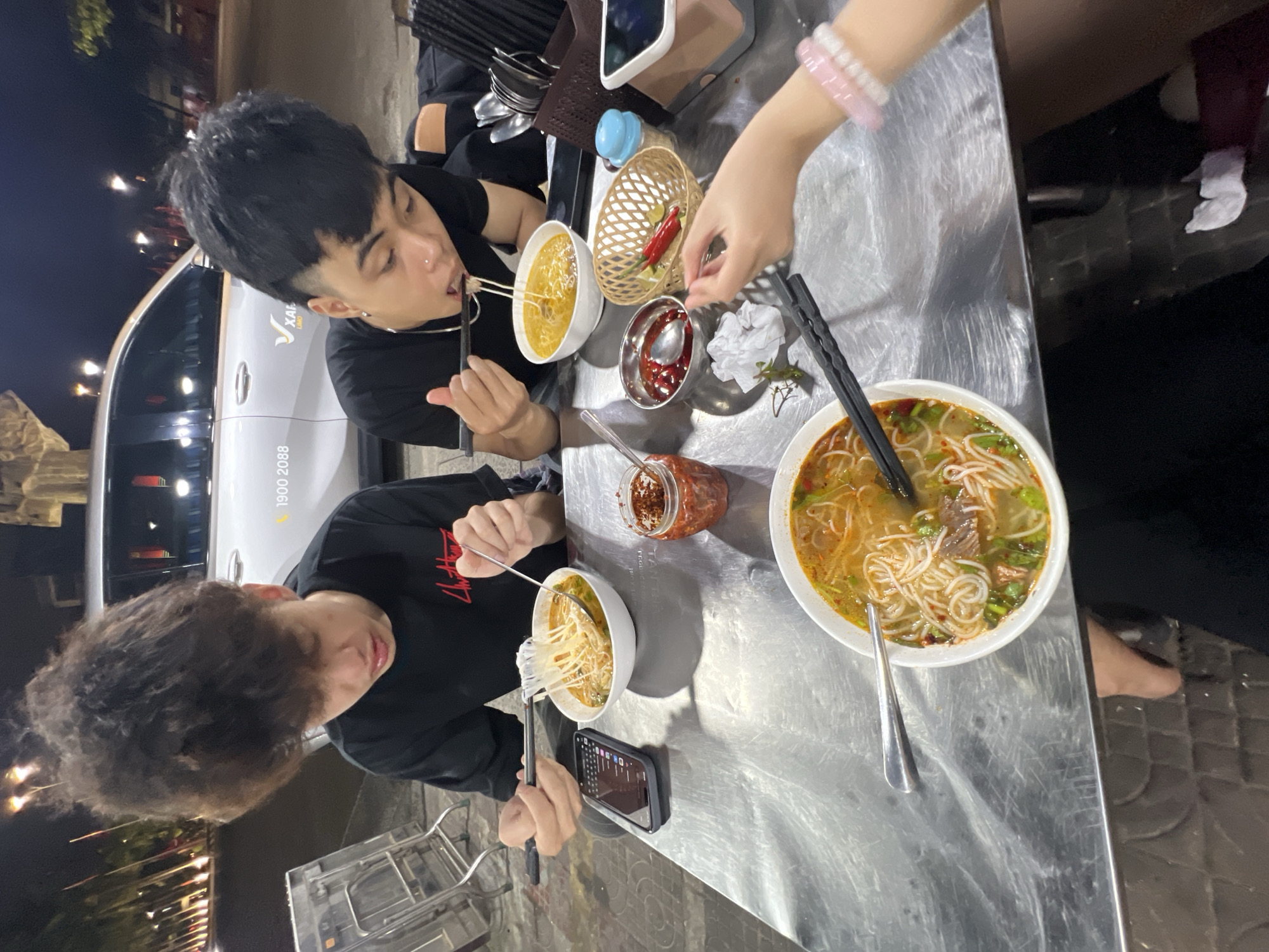 Two young men are seated at a stainless-steel table, eating Vietnamese noodle soup. A third person is visible with an arm extending into the scene to add a chili pepper to a bowl. One person has black hair and is eating from a yellow broth. The other person has curly hair and is holding a phone while eating. The table also holds a bowl of red chili peppers, a jar of chili oil, and a utensil holder. In the background, a car with the telephone number "1900 2088" is visible. This scene takes place in Đà Nẵng, Vietnam, with a restaurant visible across the street at night.