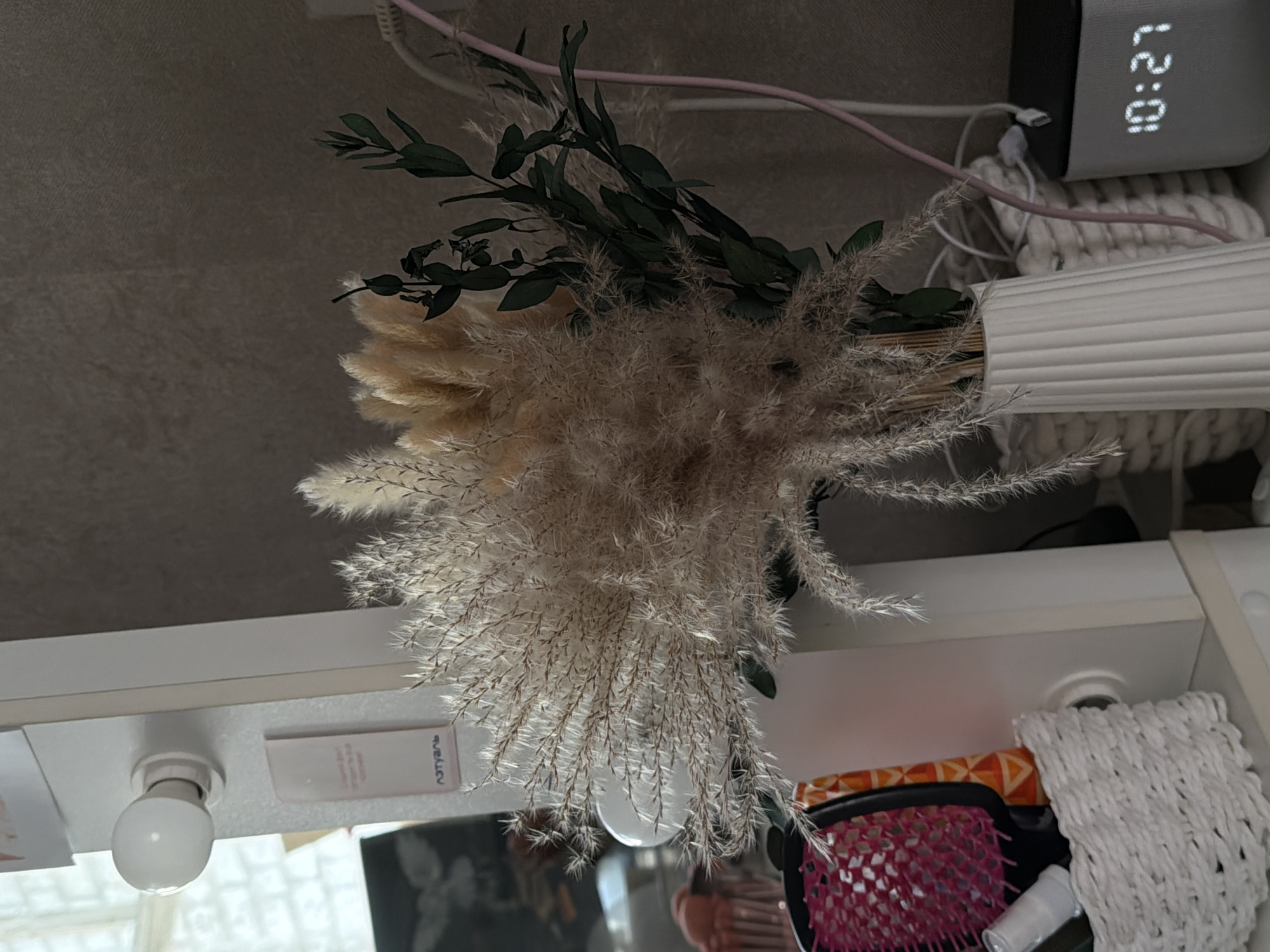 This image captures an overhead view of a styled surface, likely a shelf or dresser, in an indoor setting. The central focus is a large bouquet of dried decorative plants arranged in a white, ribbed cylindrical vase. The bouquet primarily consists of various types of feathery pampas grass and other light beige and brown dried grasses, interspersed with sprigs of dark green eucalyptus-like foliage.
To the upper right of the vase, a black digital alarm clock displays the time "10:27" in white LED digits. Several pink and white charging cables and wires are visible, tangled around the clock and a white woven object resembling a macrame mat or small basket, which is positioned beneath the clock and behind the vase.
Towards the bottom left of the frame, a white spherical lightbulb is visible, fixed to the underside of a white shelf. Next to it, on the shelf surface, lies a white rectangular object with partially visible Cyrillic text that appears to read "АЗУГТУАЛЬ" (AZUGTUAL). The bottom right corner of the image shows a partial view of personal grooming items, including a pink and black hairbrush with an orange patterned handle and a small white bottle.
No specific activity is depicted; the image presents a static still life arrangement. The time "10:27" is clearly visible on the digital clock, suggesting a morning or late morning setting. The soft, diffuse lighting enhances the delicate textures of the dried plants and the woven material. The arrangement suggests a cozy, organized personal space, potentially in a residence in Chaikovsky, Russia.