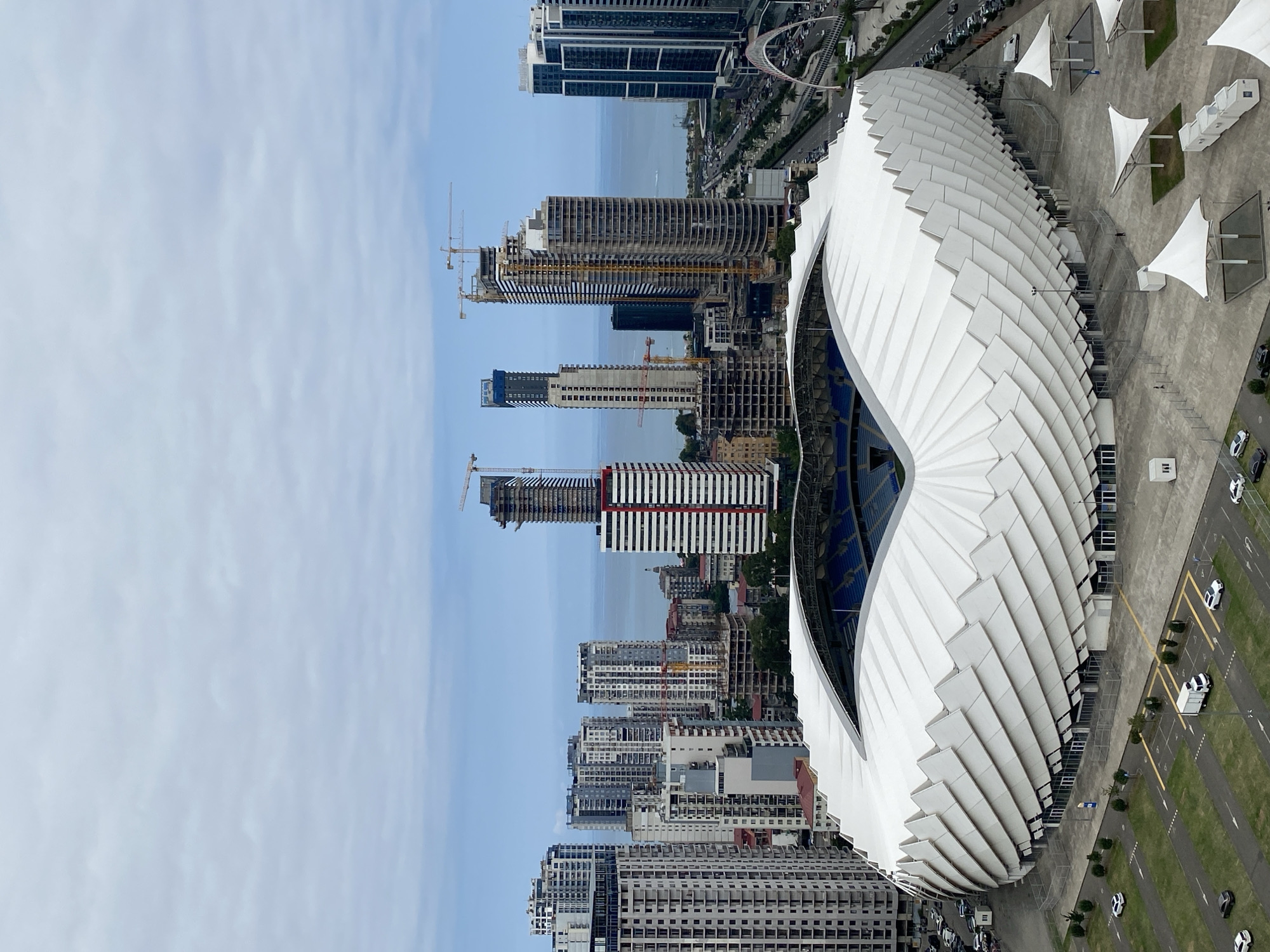 The image captures an aerial view of the Batumi Stadium in Batumi, Georgia, situated in a coastal city. The stadium, characterized by its distinctive white, wave-like roof structure, is the focal point. Surrounding the stadium, a modern cityscape unfolds, featuring numerous high-rise buildings, many of which appear to be under construction, indicated by visible cranes. A highway runs alongside the stadium, with cars and a bus visible on it. The Black Sea is discernible in the background, meeting a hazy, cloudy sky. The immediate vicinity of the stadium includes parking lots with several vehicles, including cars, a van, and what appears to be a white truck, as well as pedestrian walkways and open spaces with white canopies. The overall scene depicts a developing urban environment with significant architectural and infrastructural activity.