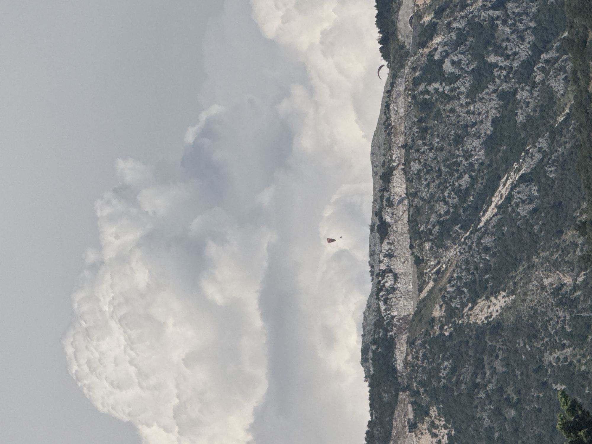 This media file captures a dramatic mountainous landscape in Boreti, Montenegro, with paragliders enjoying the activity. The scene is dominated by a towering, rugged cliff face on the right, covered in sparse vegetation and showing signs of rock erosion. To the left of the cliff, a vast expanse of cloudy sky fills the frame. The clouds are a mix of bright white, sunlit formations and darker, more diffused grey areas, suggesting a partly cloudy or overcast day.
Two individuals are visible paragliding in the sky. One paraglider, appearing as a small red and black shape, is positioned higher in the frame and closer to the cliff. Another paraglider, a small dark shape, is further to the left and slightly lower in the frame. Their presence indicates an active outdoor pursuit, likely taking advantage of the wind currents around the mountain.
The overall atmosphere is one of adventure and natural beauty. The lighting suggests it is daytime, though the cloudy conditions might mute the intensity of the sun. The sheer scale of the mountain and the vastness of the sky create a sense of awe and freedom. No visible text is present in the media file.