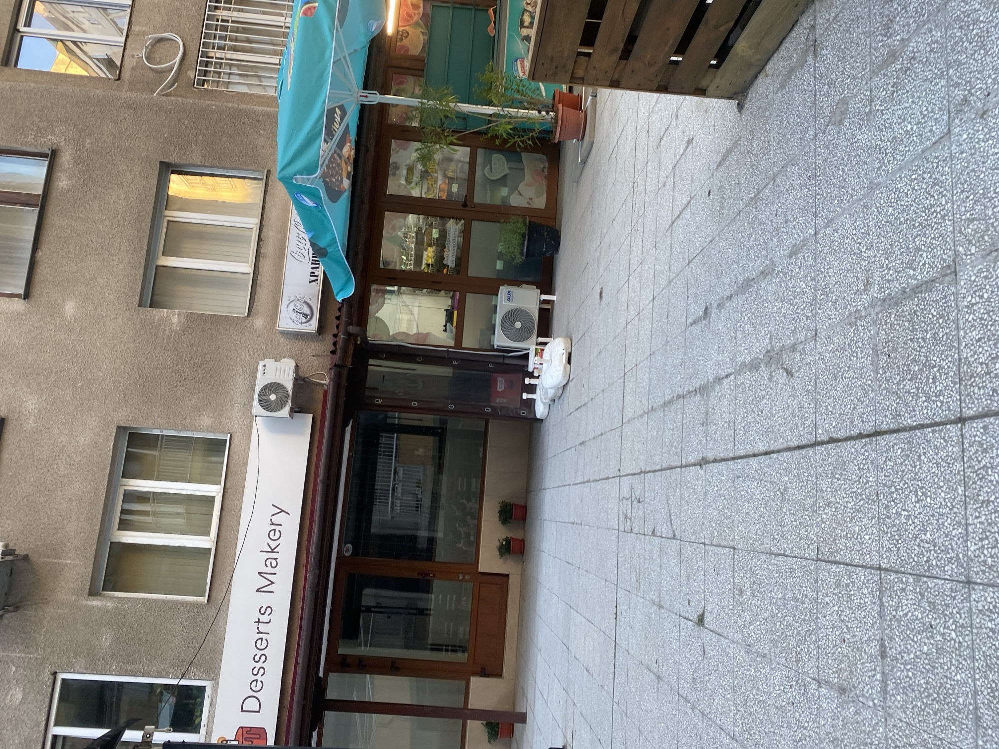 The image shows a street view in Sofia, Bulgaria. On the left is a multi-story building with a gray facade. The building has several windows with white frames. A white sign reads "Desserts Makery." An air conditioning unit is mounted on the building above the sign. To the right of the Dessert Makery is a restaurant with glass doors and windows. A blue umbrella extends from the restaurant. An air conditioning unit and several potted plants are placed in front of the restaurant. A section of the sidewalk is visible. The sidewalk is paved with gray tiles.