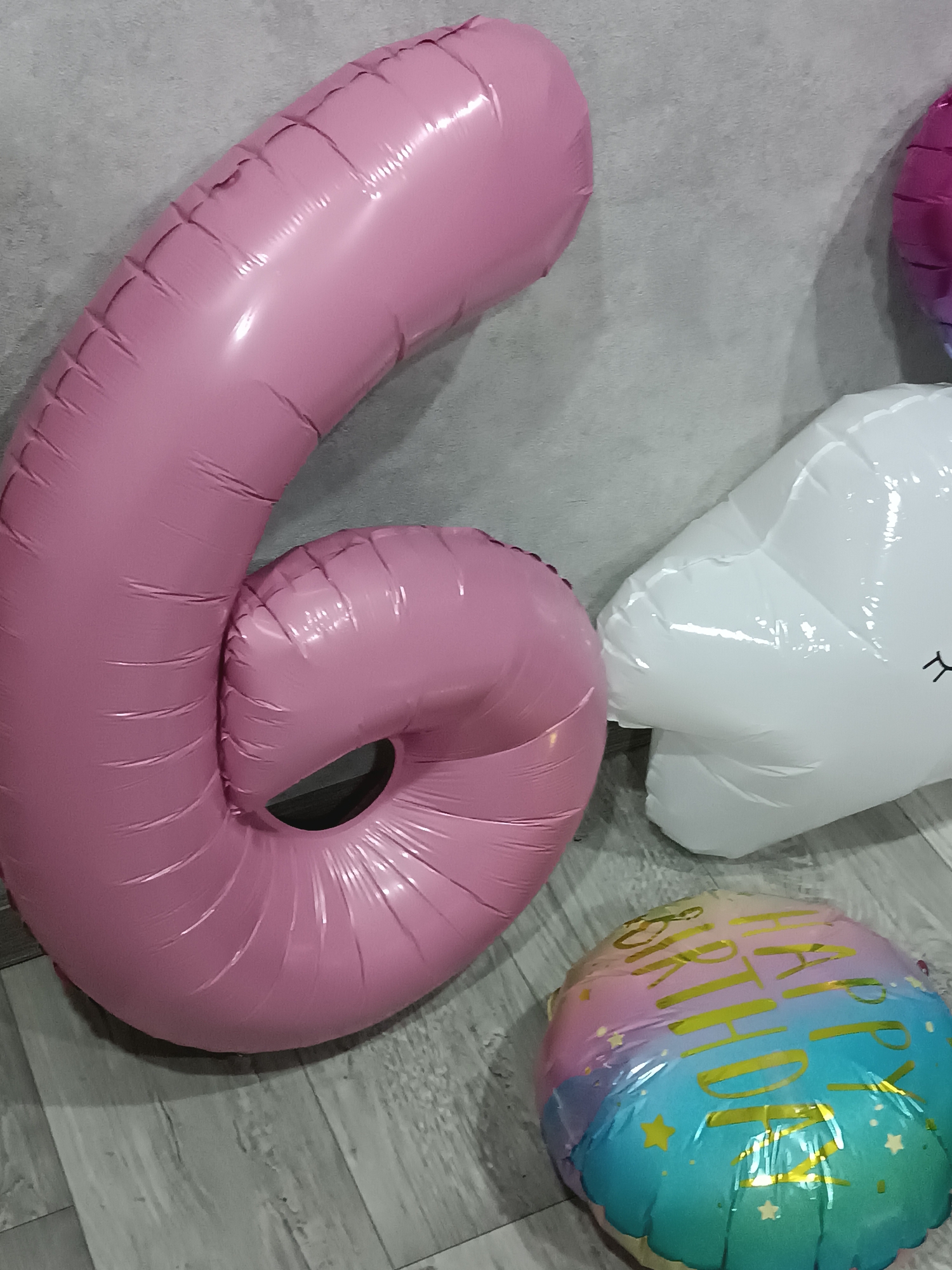 An indoor scene displays multiple inflated foil balloons. Dominating the left side is a large, pink number "6" balloon, positioned vertically against a light grey, textured wall. To the right of the pink "6" is a white, irregularly shaped foil balloon, possibly depicting a cloud, with two black, stylized eyes visible on its lower right section. Partially visible in the upper right is a magenta-colored foil balloon. On the grey wood-effect laminate floor in the lower right quadrant rests a round foil balloon with a gradient color scheme of blue, pink, and yellow, adorned with gold text that reads "HAPPY BIRTHDAY" and several small gold star graphics. All balloons exhibit surface creases and reflections from ambient light. No individuals or active interactions are present. Location: Unknown, Unknown.
