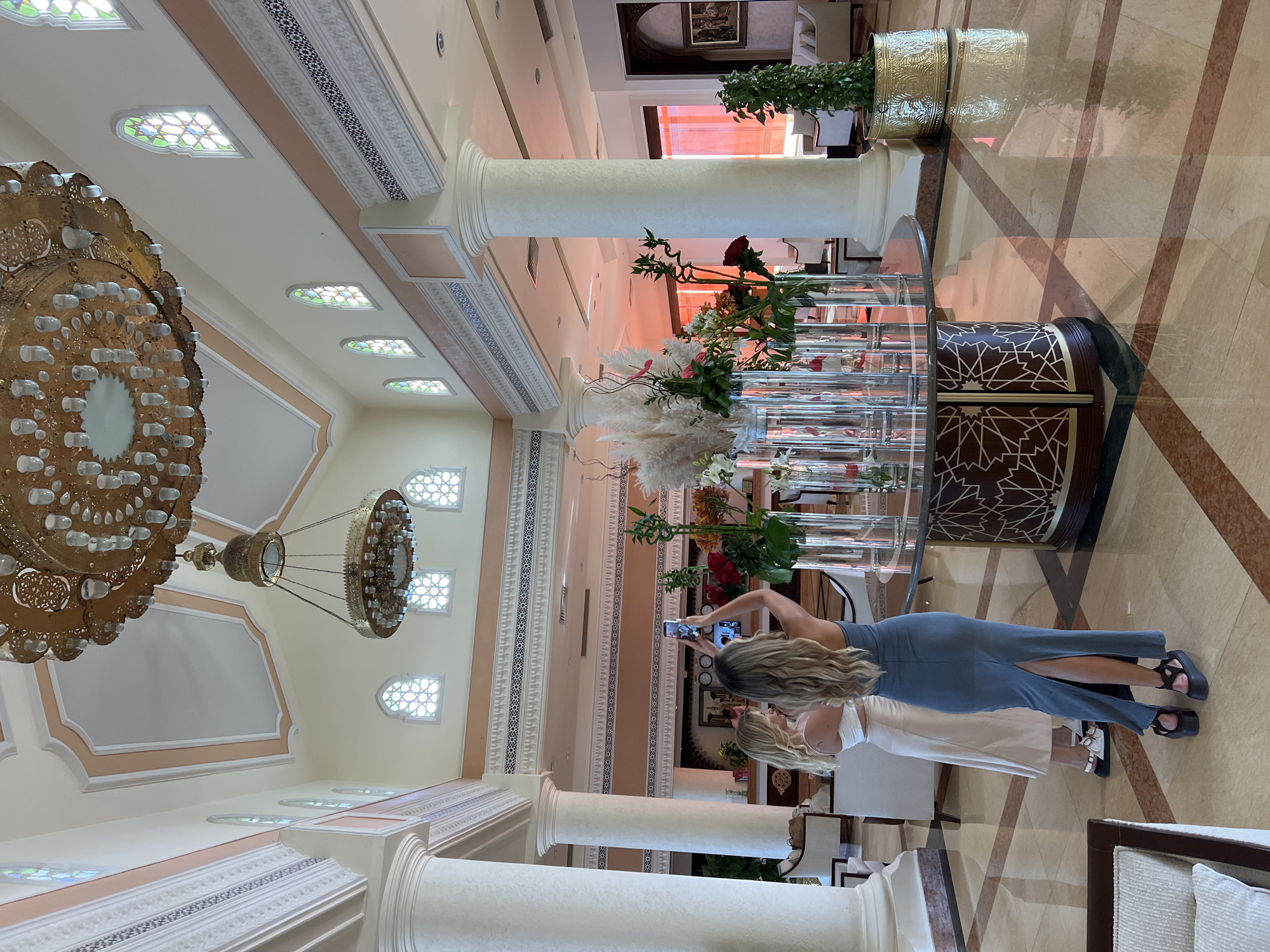 The image captures the opulent interior of a hotel lobby in Sharm El Sheikh, Egypt, characterized by its high ceilings, ornate chandeliers, and intricate architectural details. Two young women are the central human subjects. One, wearing a light-colored skirt and top, stands with her back to the camera. The other, in a long, dark blue dress with a slit, is actively taking a photograph of the lobby with her smartphone, holding it at arm's length.
The lobby features grand, cream-colored columns and a decorative frieze running along the walls, adorned with geometric patterns. Above, two large, elaborate chandeliers hang prominently, one brass and intricately detailed, the other a more modern, multi-tiered design. Stained-glass windows, framed by decorative molding, punctuate the upper walls, adding colorful light to the space.
In the foreground, a large, round table serves as a centerpiece, adorned with an extravagant floral arrangement in tall glass vases. The flowers are a mix of tall, feathery pampas grass, red roses, and other vibrant blooms. The table itself has a dark wood base with geometric inlay patterns. The flooring is a polished marble with inlaid geometric patterns in contrasting colors.
In the background, a reception desk is visible, with a clock displaying multiple time zones. Seating areas with plush furniture are scattered throughout the lobby. The overall ambiance suggests a luxurious and well-maintained establishment. The warm lighting, reflecting off the polished surfaces, creates a welcoming and sophisticated atmosphere, indicative of a high-end resort or hotel in a tourist destination like Sharm El Sheikh.