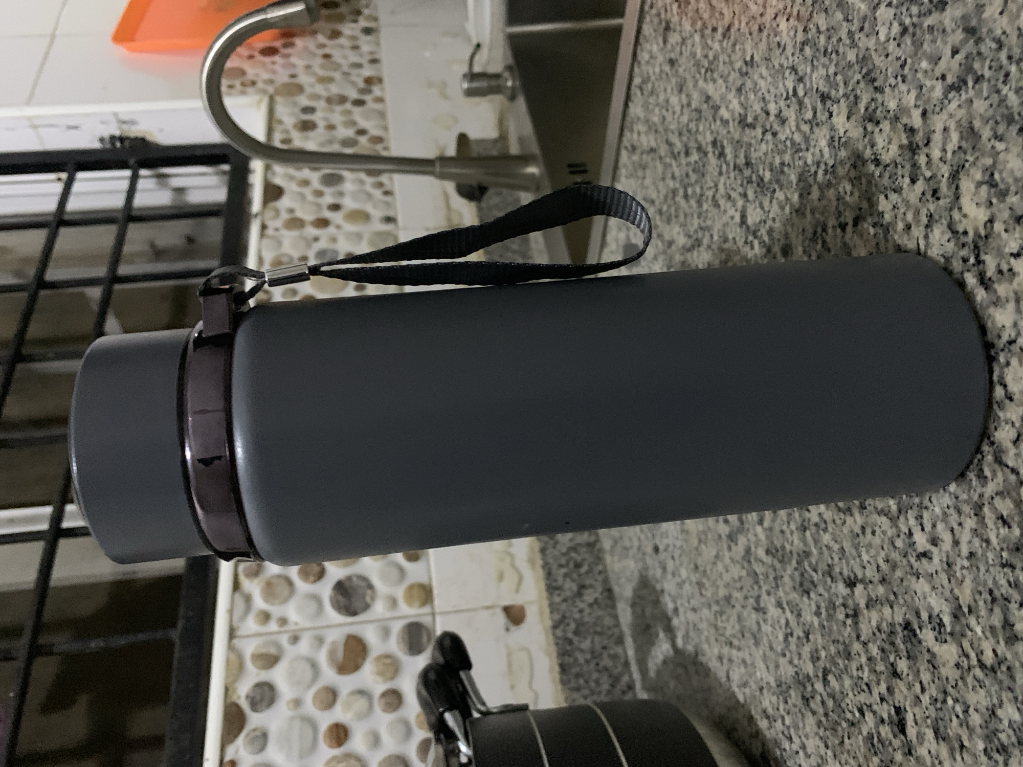 An indoor photograph, taken in a kitchen or utility area in Gwarinpa, Nigeria, displays a gray, insulated water bottle or flask lying horizontally on a granite countertop. The bottle is cylindrical with a matte finish and features a black fabric strap attached to a dark metallic band near its cap. The strap has a finely ribbed texture, visible in close-up. A minor scuff mark is visible on the metallic band around the bottle's neck.
In the background, behind the bottle, a stainless steel kitchen faucet with a curved spout is prominent, extending from a sink area which is partially visible. An orange plastic tray can be seen under the faucet, suggesting a dish-washing or drying area. The wall behind the sink is covered with small, irregularly shaped mosaic tiles in shades of brown, beige, and white. To the far left, a black metal grate structure is partially visible, likely a window or security bars. At the bottom of the frame, another black object, possibly a part of a stove, kettle, or another appliance, is dimly seen. The overall lighting suggests an indoor setting, likely with artificial light. There is no discernible text on any of the visible objects. No specific activity is taking place; the bottle simply rests on the counter.