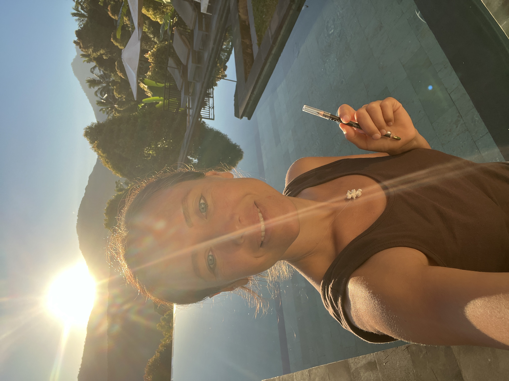 A woman with brown hair and blue eyes is taking a selfie by a swimming pool in Busungbiu, Indonesia. She is wearing a brown tank top and a white flower pendant necklace. She is holding a pencil in her right hand. The sun is setting behind mountains in the background, casting a warm glow over the scene. The swimming pool reflects the sky and surrounding lush greenery. There are lounge chairs and umbrellas visible by the pool, suggesting a resort or hotel setting. The overall atmosphere is peaceful and serene.