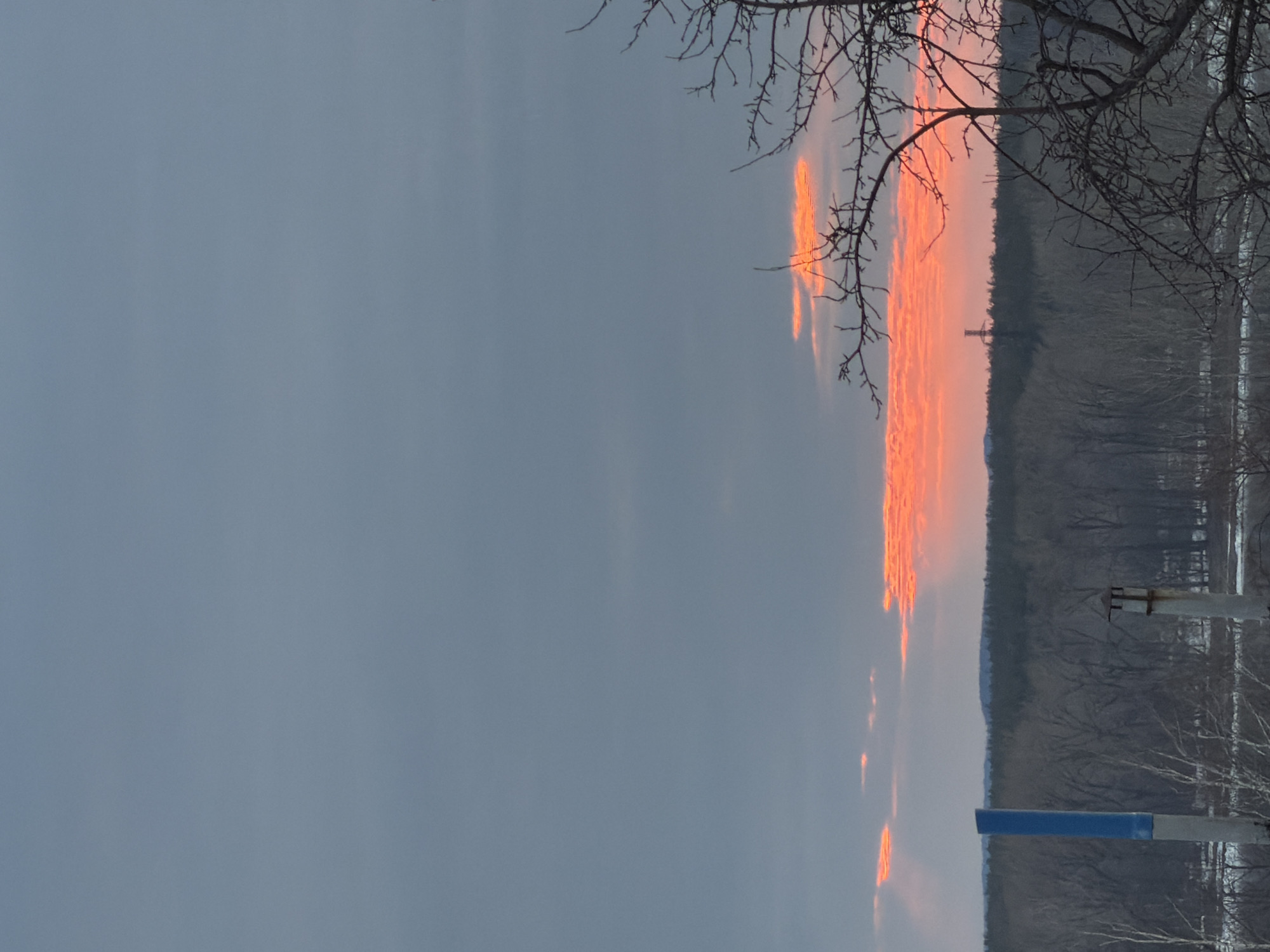 The image captures a winter scene in Chaikovsky, Russia, at dusk or dawn. The sky is predominantly a dull grey, with a distinct line of orange-red light indicating the horizon. Bare tree branches extend from the top-right corner of the frame. A line of trees and vegetation form the backdrop, with what appears to be a structure or tower visible in the distance. In the foreground, a blue rectangular sign atop a pole can be seen. The overall impression is of a cold, subdued landscape on the outskirts of the city.