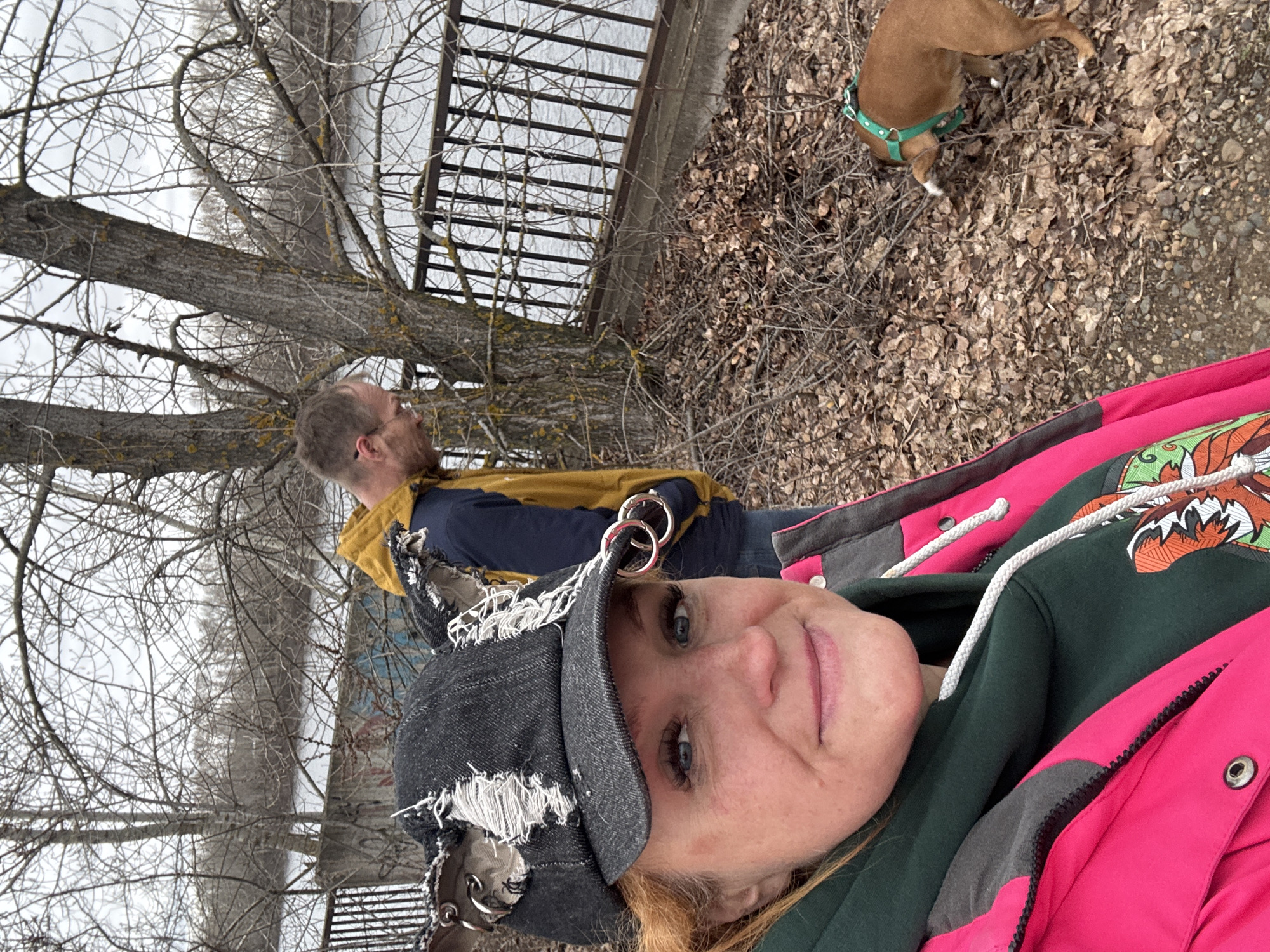 The image captures an outdoor scene, likely a riverside or park area in Chaikovsky, Russia, characterized by bare trees and an overcast sky, suggesting late autumn, winter, or early spring.
In the foreground, a woman takes a selfie, looking directly at the camera with a slight smile. She has light-colored hair and wears a distressed dark denim baseball cap adorned with metal rings and frayed fabric. She is dressed in a dark green hoodie featuring a distinctive, colorful graphic resembling a fox or other animal, visible on the chest, layered under a bright pink and gray jacket with a prominent zipper and drawstrings.
Behind her, a man with short hair and a beard is seen in profile, looking upwards and to his right. He wears a yellow jacket over a dark blue long-sleeved shirt. Further back and to the right, a small to medium-sized brown dog with a bright green harness is sniffing the ground.
The ground is covered with a thick layer of dry, brown fallen leaves and dirt. In the background, numerous deciduous trees with dark, gnarled trunks and intricate bare branches dominate the left side of the frame, reaching towards a uniformly gray, overcast sky. A dark metal fence runs along what appears to be the edge of a body of water, possibly a river or lake, visible behind the man and the dog.
The overall atmosphere is quiet and casual, depicting a group enjoying an outdoor walk. There is no visible text in the image.
