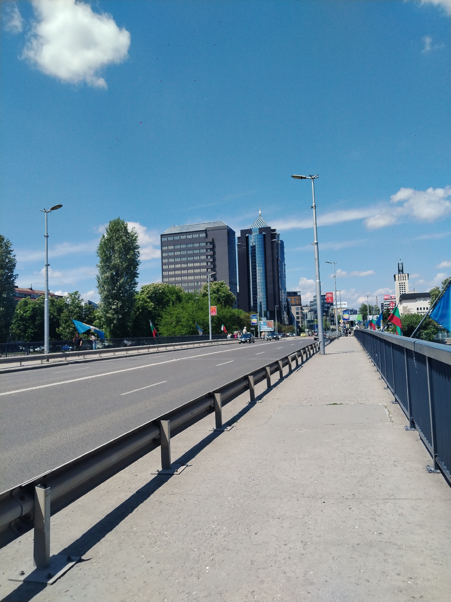 The image shows a view from a bridge in Plovdiv, Bulgaria. The bridge has a metal guardrail separating the sidewalk from the road. A grey metal barrier divides the road from the sidewalk. A black office building with a glass tower is visible, along with a building with a tower. Streetlights line the bridge. Bulgarian flags are attached to the guardrail. Cars are visible on the road, and people appear to be walking on the sidewalk on the far side of the bridge. The sky is blue with white clouds.