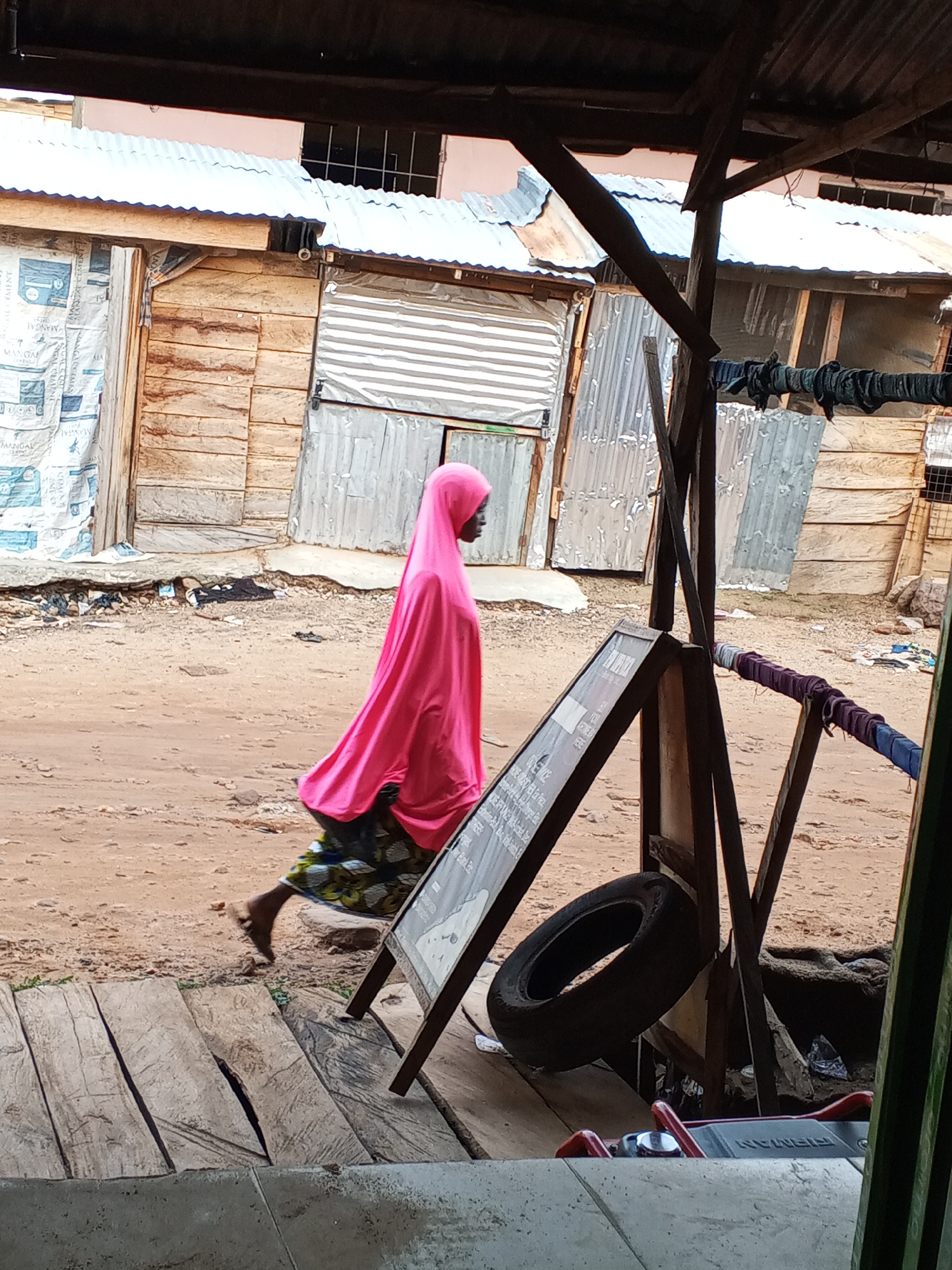 The image depicts a street scene in Lokoja, Nigeria. A woman wearing a bright pink headscarf and a dark, patterned skirt is walking along a dirt road. The road is lined with makeshift structures constructed of wood and corrugated metal. Laundry hangs on a line strung between two of the structures. In the foreground, a wooden sign and a tire lean against a wooden post. The scene is viewed from under a shelter, possibly the eaves of a building.