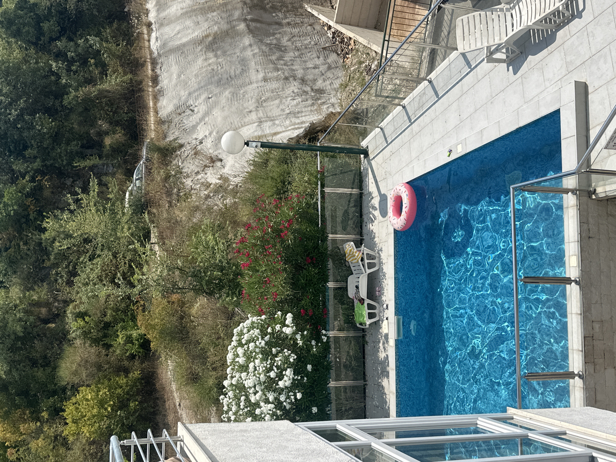 The image depicts a private outdoor swimming pool area, likely part of a villa or residential property, located in Herceg Novi, Montenegro. The pool is rectangular and filled with bright blue, sunlit water, with shimmering light patterns on its surface. A pink inflatable ring float is visible in the water. The pool area is paved with light-colored square tiles.
Surrounding the pool, there is a white plastic lounge chair with a yellow and white striped towel on it, and another white lounge chair with a green towel and some slippers. Behind the pool, there are lush green bushes, including a large cluster of white flowers and a smaller cluster of red flowers. A metal fence with glass panels encloses the pool area.
In the background, a steep, rocky hillside is visible, with dense green trees and vegetation covering the upper parts. A dirt road runs along the hillside, and a dark-colored car is parked on this road. A modern street lamp with a spherical white globe is positioned on the paved area near the pool. The overall scene conveys a sense of a sunny, relaxed atmosphere in a natural, hilly environment.