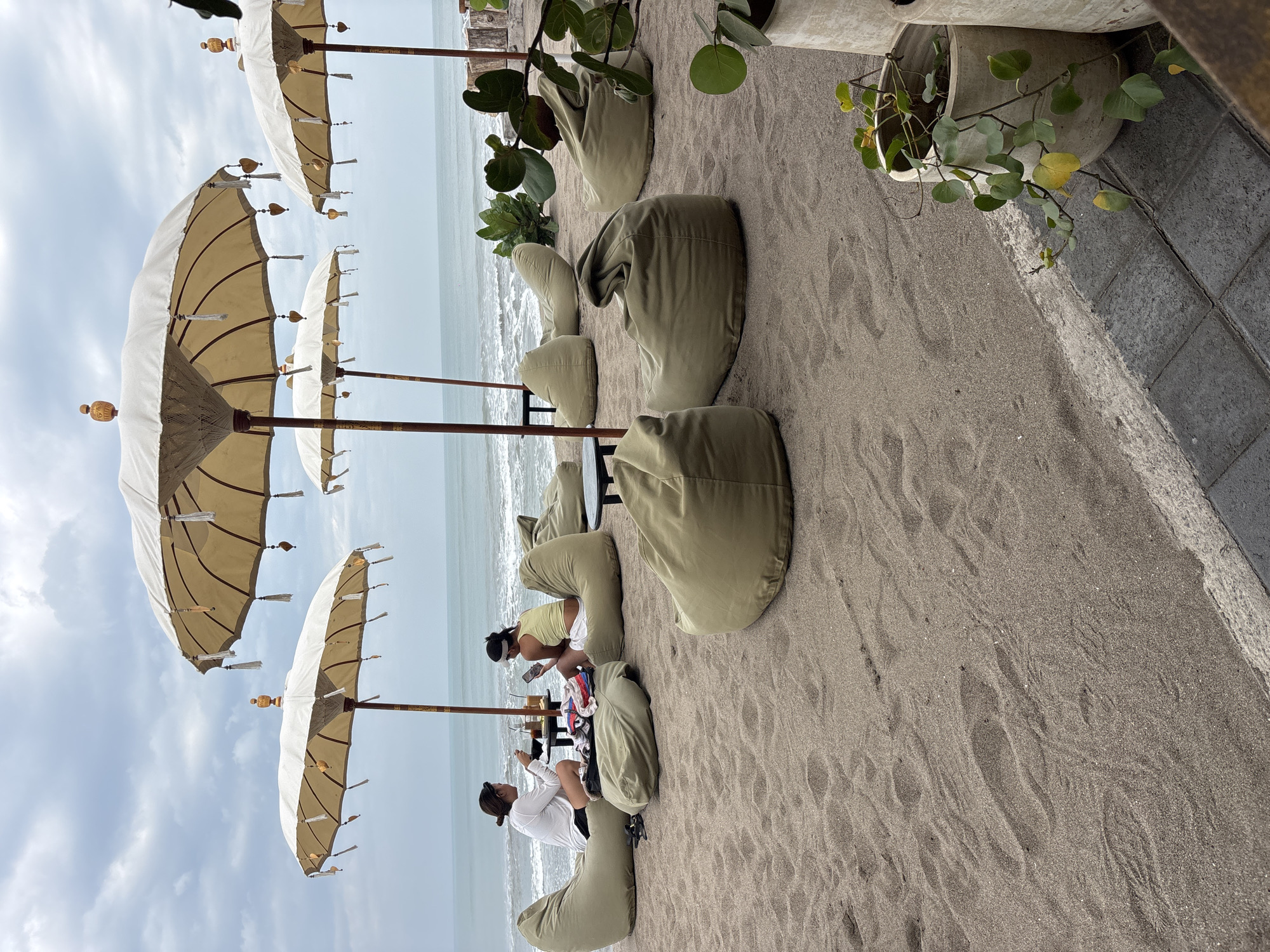 The scene is a beachfront area in North Kuta, Indonesia, featuring a relaxed outdoor seating arrangement. Several large, pale beige beach umbrellas with decorative tassels and heart-shaped charms are erected on the sand, providing shade. In the foreground and midground, numerous large, light olive green beanbag chairs are scattered across the sandy surface. Two women are seated on beanbag chairs at a small black table in the midground. One woman, wearing a white long-sleeved shirt and black shorts, is looking down at her food and drink. The other woman, in a yellow tank top and white shorts, is looking at her phone. Both women have their hair tied back. The ocean is visible in the background, with calm waves and a cloudy sky overhead. To the right of the frame, a grey paved pathway runs along the edge of the sand, bordered by a low wall. Potted plants with green leaves are placed near the pathway. The sand has visible footprints.