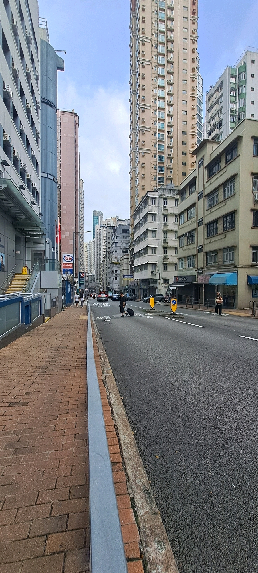 The image depicts a street scene in a dense urban environment, likely Hong Kong, given the architectural style and the presence of a red and white Esso sign. The perspective is from a sidewalk looking towards a street lined with tall buildings. The sidewalk is paved with red bricks and has a concrete curb. The street is paved with asphalt and appears to be a multi-lane road with traffic.
In the foreground, a person is seen walking across the street, pulling a rolling suitcase. This individual is dressed in dark clothing. Further down the street, another person is visible on the sidewalk. Several vehicles are on the road, including cars and possibly a bus in the distance.
The buildings are of various heights and architectural styles, characteristic of Hong Kong's urban landscape. Some are modern high-rises with numerous windows and air conditioning units, while others appear older, with more traditional facades. There are shops and businesses at street level, indicated by signage like "Pizza Hut." The sky is overcast, suggesting a cloudy day. The overall impression is a busy, commercial street in a densely populated area.