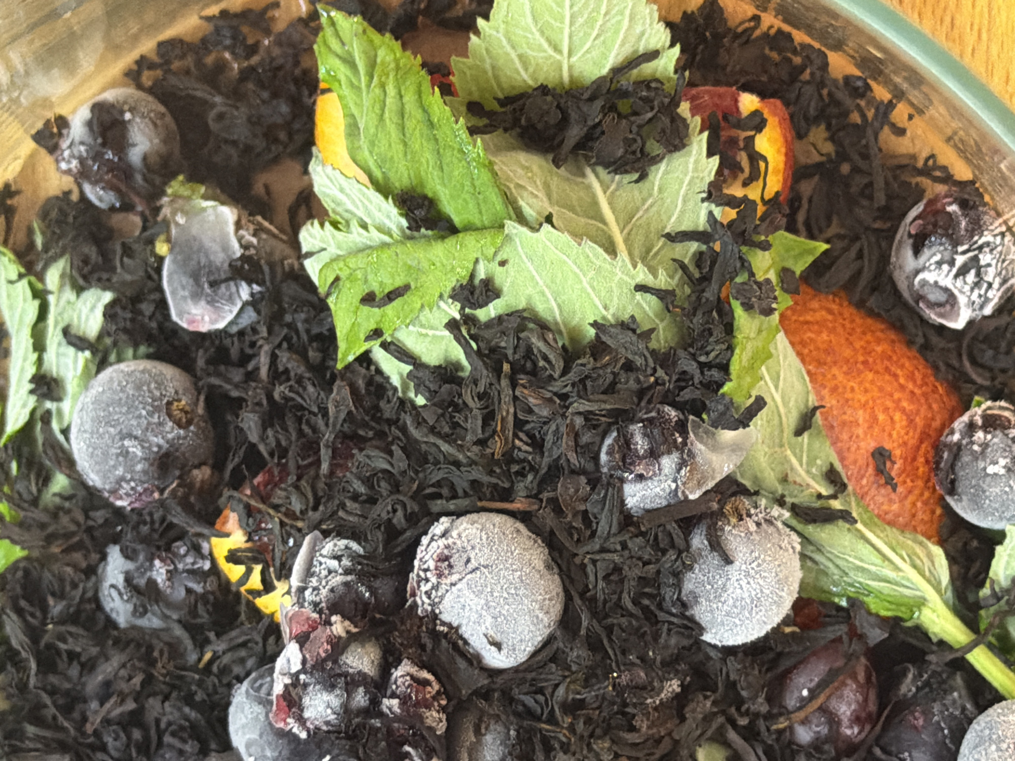 The image shows a close-up view of dried tea leaves mixed with several frozen berries, mint leaves, and slices of orange. The berries appear to be blueberries and possibly some other dark fruit, all coated in a layer of frost. The mint leaves are a vibrant green and some appear to be slightly wilted. The orange slices have a bright orange rind and fleshy interior. The tea leaves are dark and appear to be of the loose-leaf variety. The composition suggests this mixture is intended for brewing. There is no indication of a specific location in the city of Шмидтово, Russia, or any people or actions. The scene is static and focuses solely on the ingredients of the tea mixture.