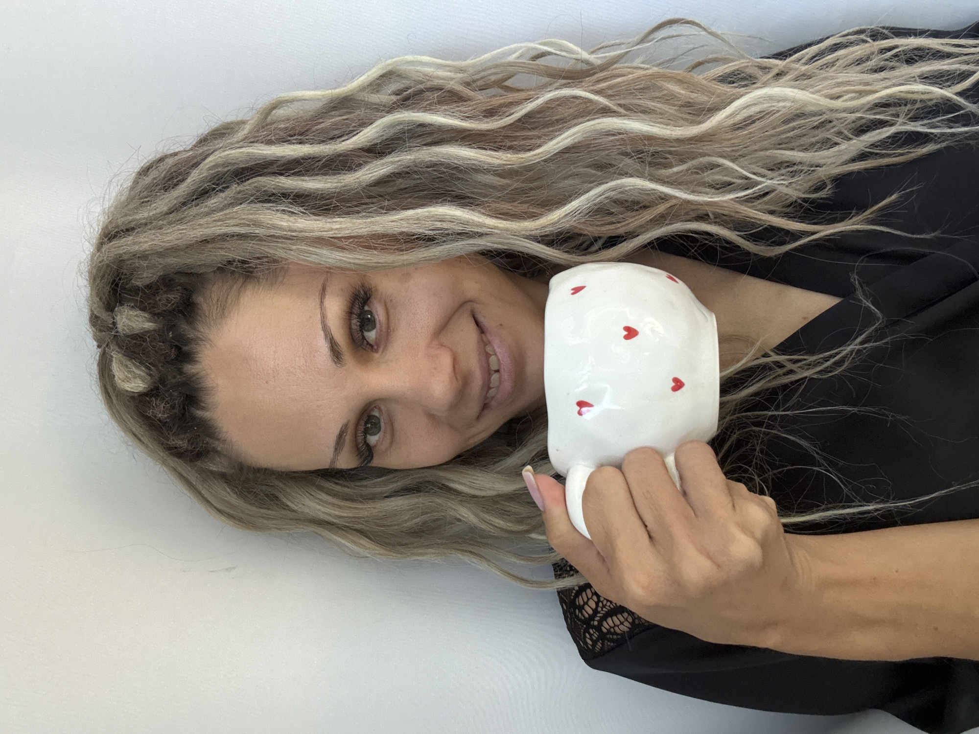 A medium shot shows a woman with blonde dreadlocks, holding a white mug with red hearts and looking at the camera. She is wearing a black robe. The background is a plain white wall. The location is Rusanovo, Russia.