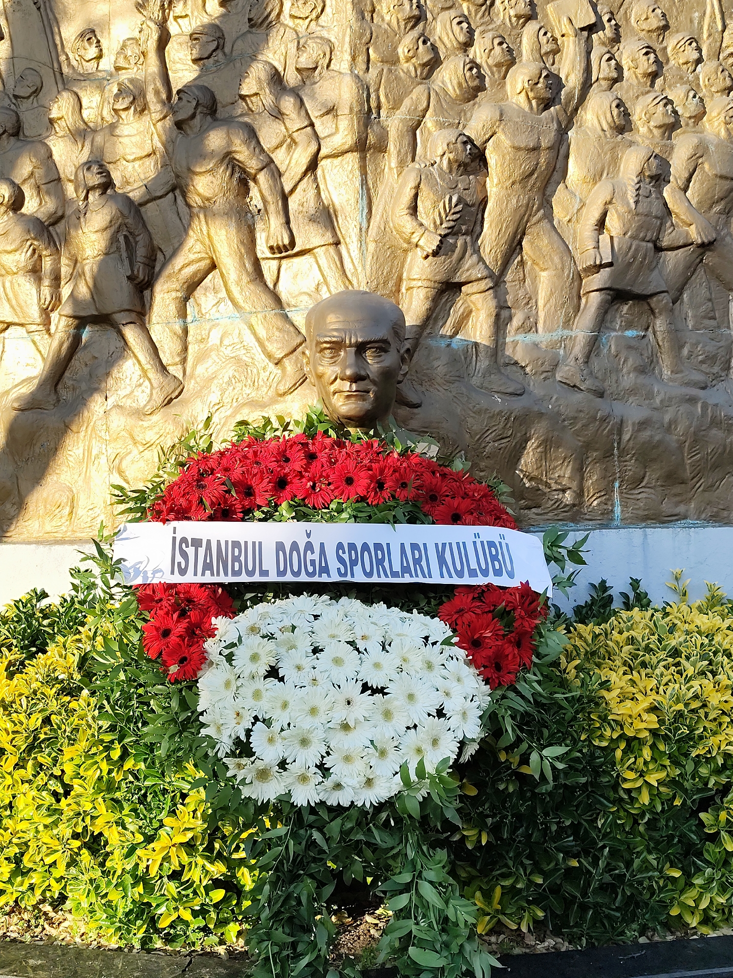 The image shows a bronze bust of Mustafa Kemal Atatürk, the founder of the Republic of Turkey, positioned in front of a bronze relief depicting a crowd of people. A wreath is placed around the base of the bust, comprised of red and white flowers, with a white banner reading "ISTANBUL DOGA SPORLARI KULUBU". The environment is likely a public square in Istanbul, Turkey. The time of day appears to be daytime, based on the lighting.